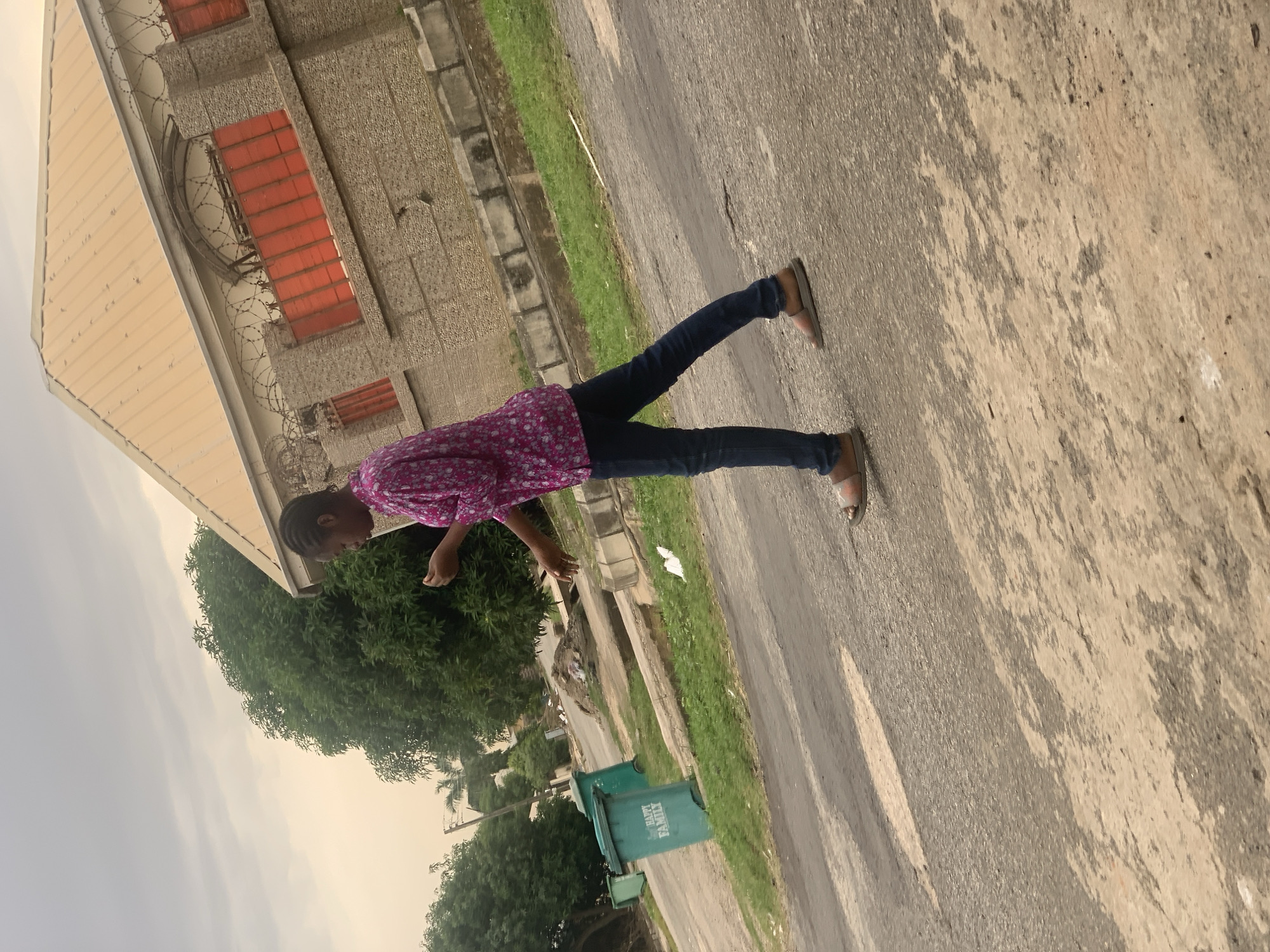 A young Black woman in a pink floral shirt and dark blue jeans walks on a cracked asphalt road in Abuja, Nigeria. She wears brown sandals. To her left, two green trash bins with "HAPPY FAMILY" written on them sit near the road. In the background, a tan building with red-paned windows and barbed wire atop a stone wall is visible. Trees and a cloudy sky complete the scene.