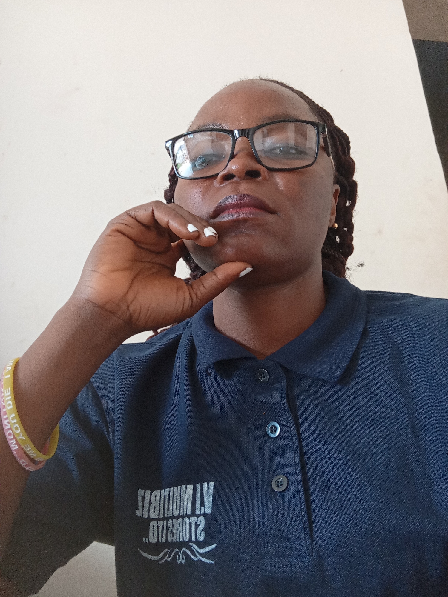 A medium shot shows a person wearing glasses and a dark blue polo shirt. The person is resting their chin on their left hand, which is partially obscuring their face. The polo shirt has a logo with white lettering that appears to be reversed. Two bracelets, one yellow and one pink, are visible on the person's left wrist. The background is a plain, light-colored wall. The image appears to have been taken indoors, possibly in Jalingo, Nigeria, based on the logo on the shirt.