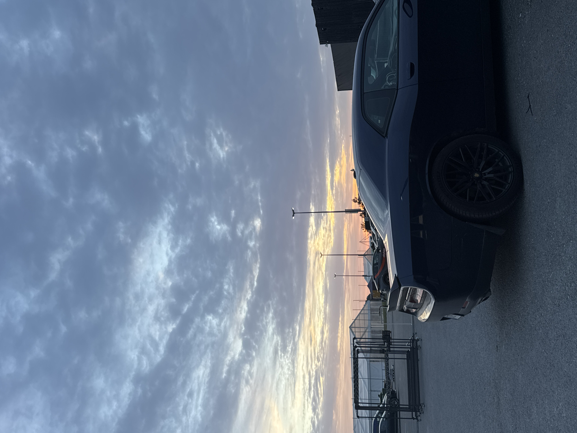 A dark blue Dodge Challenger is parked in an outdoor lot at dusk, with a dramatic cloudy sky overhead. The car's rear quarter panel and wheel are prominently featured in the foreground. In the background, several large white tent structures are visible, suggesting a temporary event or market. The ground is asphalt. The image appears to be taken in Tallinn, Estonia, given the presence of a car with a license plate that matches Estonian registration plates. The lighting indicates either early morning or late evening.