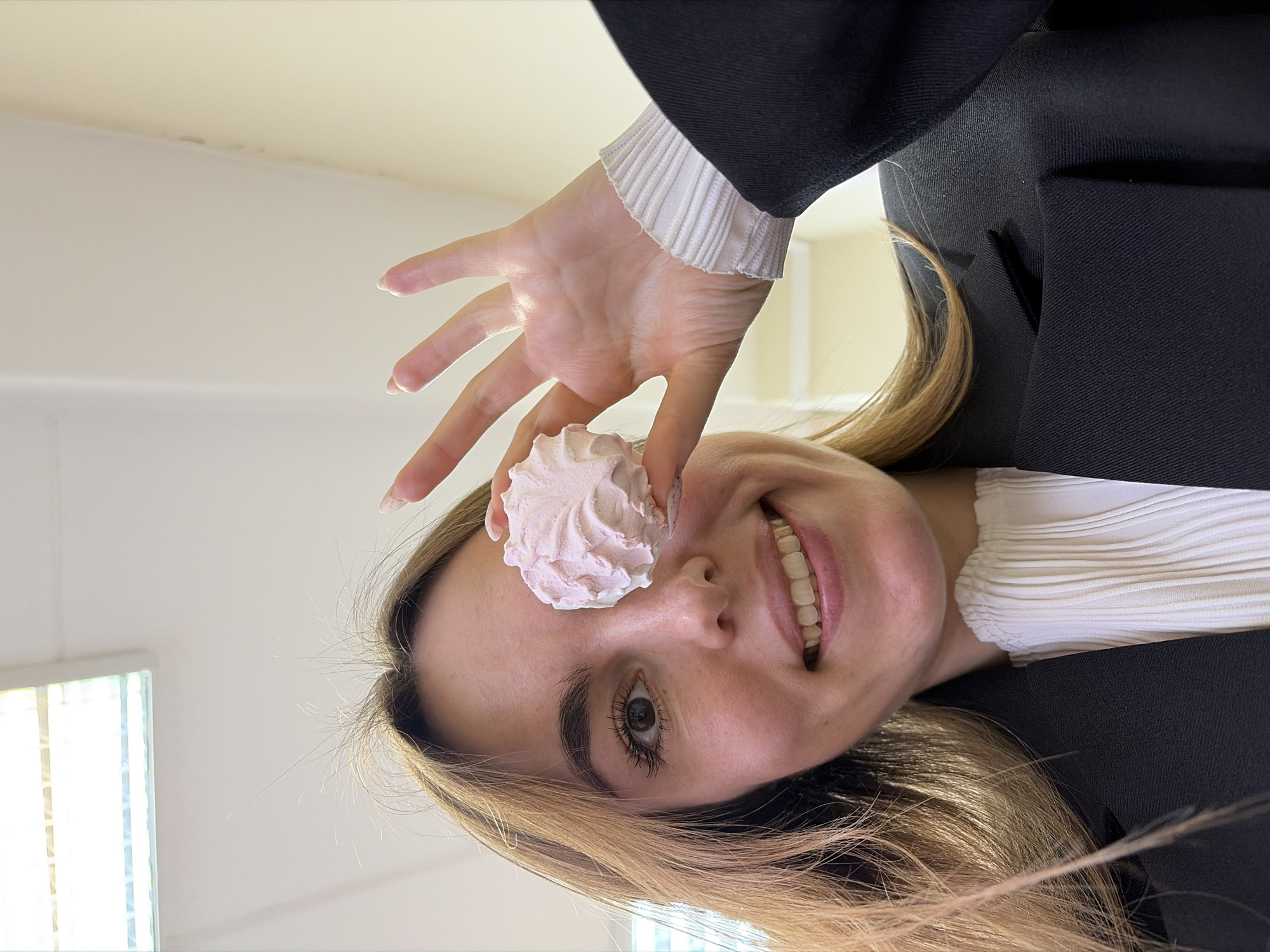 A fair-skinned woman is holding a pink meringue over her right eye. She is smiling, and her teeth are visible. She is wearing a white pleated shirt under a black blazer, and the sleeve of the shirt is visible at the wrist. The background appears to be a room with white walls, and a window is visible. The location in the city of Izhevsk, Russia, cannot be confirmed from this image.