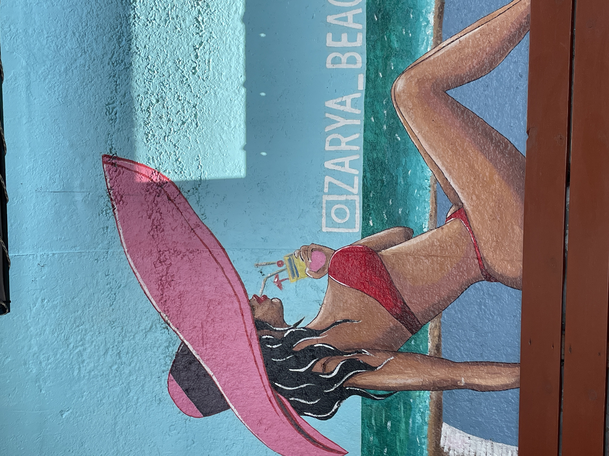The mural depicts a woman in a red bikini lounging on a beach chair, holding a cocktail, under a large pink sun hat. She is positioned against a bright blue wall, with a depiction of the sea and the text "ZARYA_BEACH" visible. This mural is located in the city of Sochi, Russia. The style is illustrative, with bold colors and defined lines. The woman is shown in profile, with her eyes closed, suggesting relaxation. The overall scene conveys a summery, vacation atmosphere.
