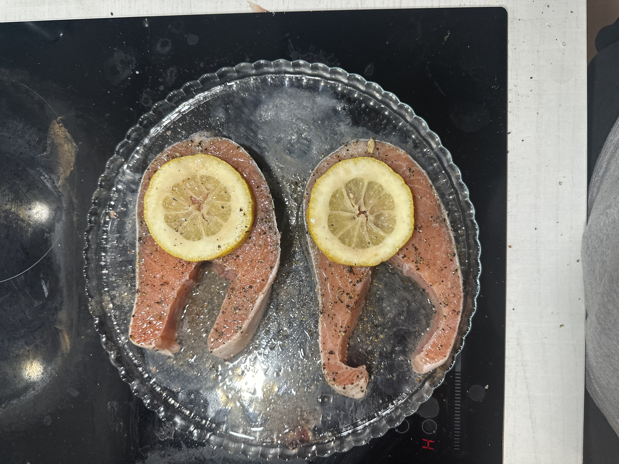 Two raw salmon steaks, seasoned with black pepper and other spices, are centered in a clear, round glass baking dish with a scalloped edge. Each salmon steak has a thin slice of lemon placed on top of it. The dish appears to contain some liquid, likely oil or marinade, reflecting light.
The scene is set on a black induction stovetop, which shows some smudges and reflections. To the right of the stovetop is a light-colored, possibly white or light grey, countertop. A small portion of a grey garment or fabric is visible in the bottom right corner, partially obscuring the countertop.
On the control panel area of the induction stovetop, a small red illuminated "H" or "4" is visible, typically indicating a hot surface or an active hob. The image captures a moment of food preparation in what appears to be a kitchen. No people or specific weather conditions are visible, and no overt emotions are expressed. The overall impression is one of preparing a meal.