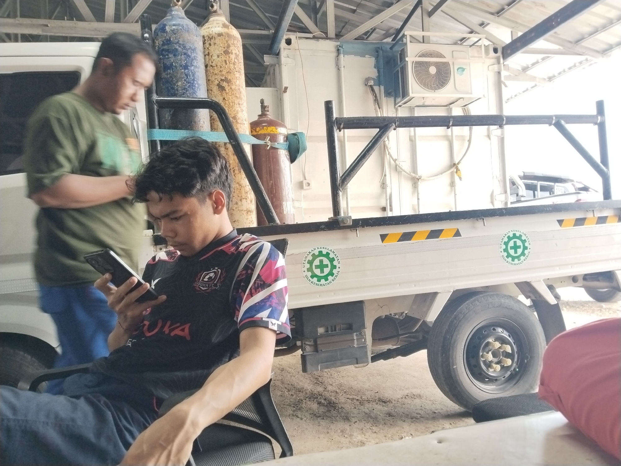 In Watuagung, Indonesia, a young man in a black and colorful soccer jersey sits and looks at his phone, his left arm resting on the armrest of a black chair. He is wearing blue jeans. To his left, another man in a green t-shirt and blue jeans stands near a white vehicle, looking down. In the background, several large gas cylinders are visible, some yellow and rusted, others blue and weathered. A white truck bed is also prominent, with a metal frame structure attached. An air conditioning unit is mounted on the side of the truck bed. The environment appears to be an outdoor industrial or storage area, with a dirt or gravel ground and a corrugated metal roof overhead. A green circular safety emblem with a cross is visible on the side of the truck bed.