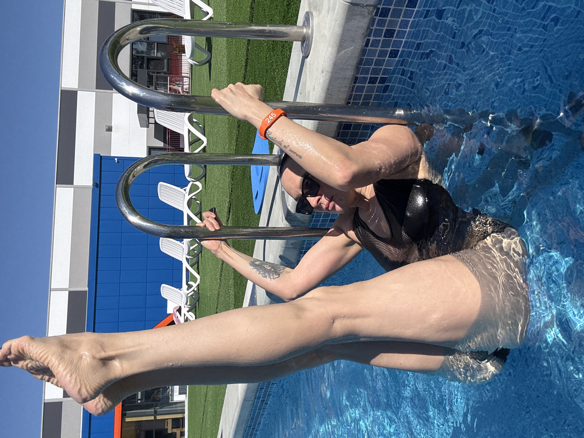 A woman in a black bathing suit and sunglasses is standing in a swimming pool, holding onto a metal ladder. She is wearing an orange wristband with the number "245" on it. Her legs are extended into the pool. The pool is lined with blue tiles and the water is clear. In the background, there is a building with blue and grey panels, white lounge chairs, and green grass. The location is potentially in Votkinsk, Russia.