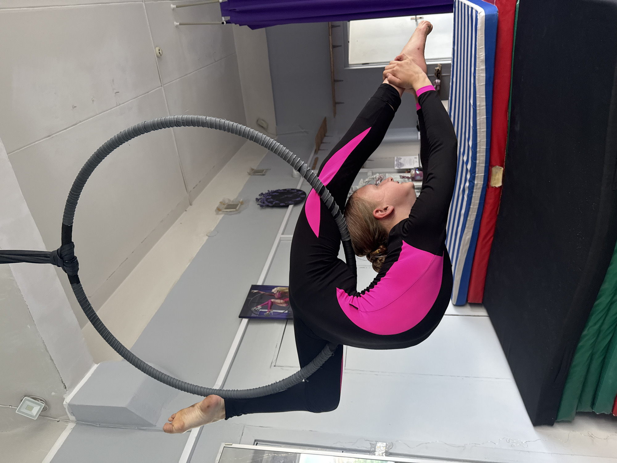 A young person with blonde hair pulled back in a ponytail, wearing a black and bright pink long-sleeved leotard, is suspended mid-air inside a gray aerial hoop. Their body is contorted into a challenging pose, with their legs extended and their feet held by their hands. The hoop is suspended from the ceiling by black fabric straps. The background shows a room with light gray walls, a white ceiling, and several stacked mats on the floor. The mats are black, green, red, and blue with white stripes. There is also a purple curtain and a poster on the wall. The lighting appears to be from overhead fixtures. The scene is consistent with a gymnastics or aerial arts training facility. The image was taken in Dnipro, Ukraine.