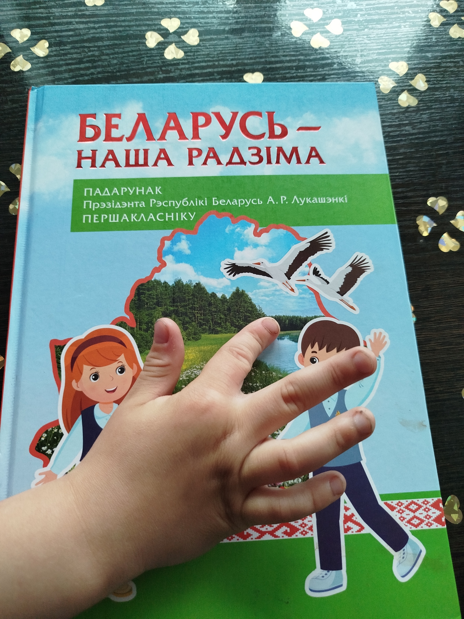 A child's hand with visible fingernails is resting on a light blue book with red and green text. The book cover features a landscape illustration with two storks flying in a blue sky above a green field and a body of water. Two cartoon characters, a girl and a boy, are depicted on the cover, dressed in casual attire. A decorative border with a red and white pattern runs along the bottom of the book. The book is placed on a dark wooden surface scattered with gold heart-shaped confetti. The exact location in Kryvaselski Sel'ski Sovet, Belarus, is not discernible from the image.