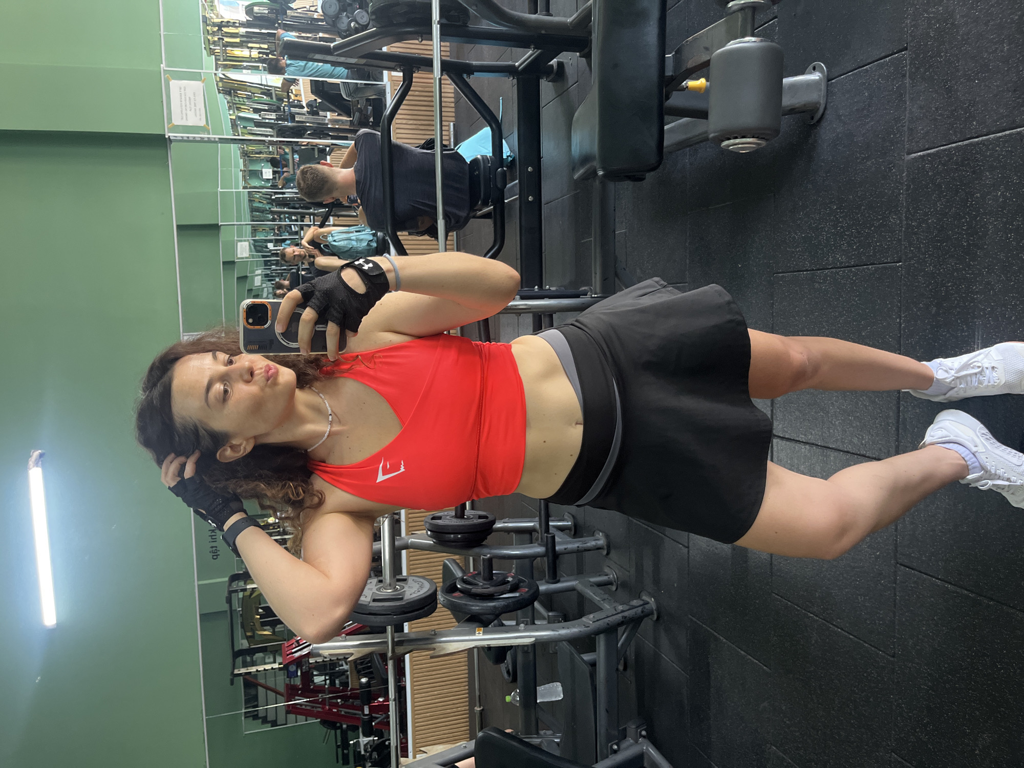 A young adult woman with curly brown hair is taking a mirror selfie in a gym. She is wearing a red sports bra and a black skirt, and white sneakers. She has on black workout gloves. She is holding her phone in her right hand, and her left hand is touching her hair. She is looking at the camera with her lips pursed. In the background, there are weightlifting machines and other gym equipment. There are also other people in the gym, some working out and some sitting. The gym appears to be clean and well-lit. The location is the Nova Fitness & Yoga gym in Phan Thiết, Vietnam.