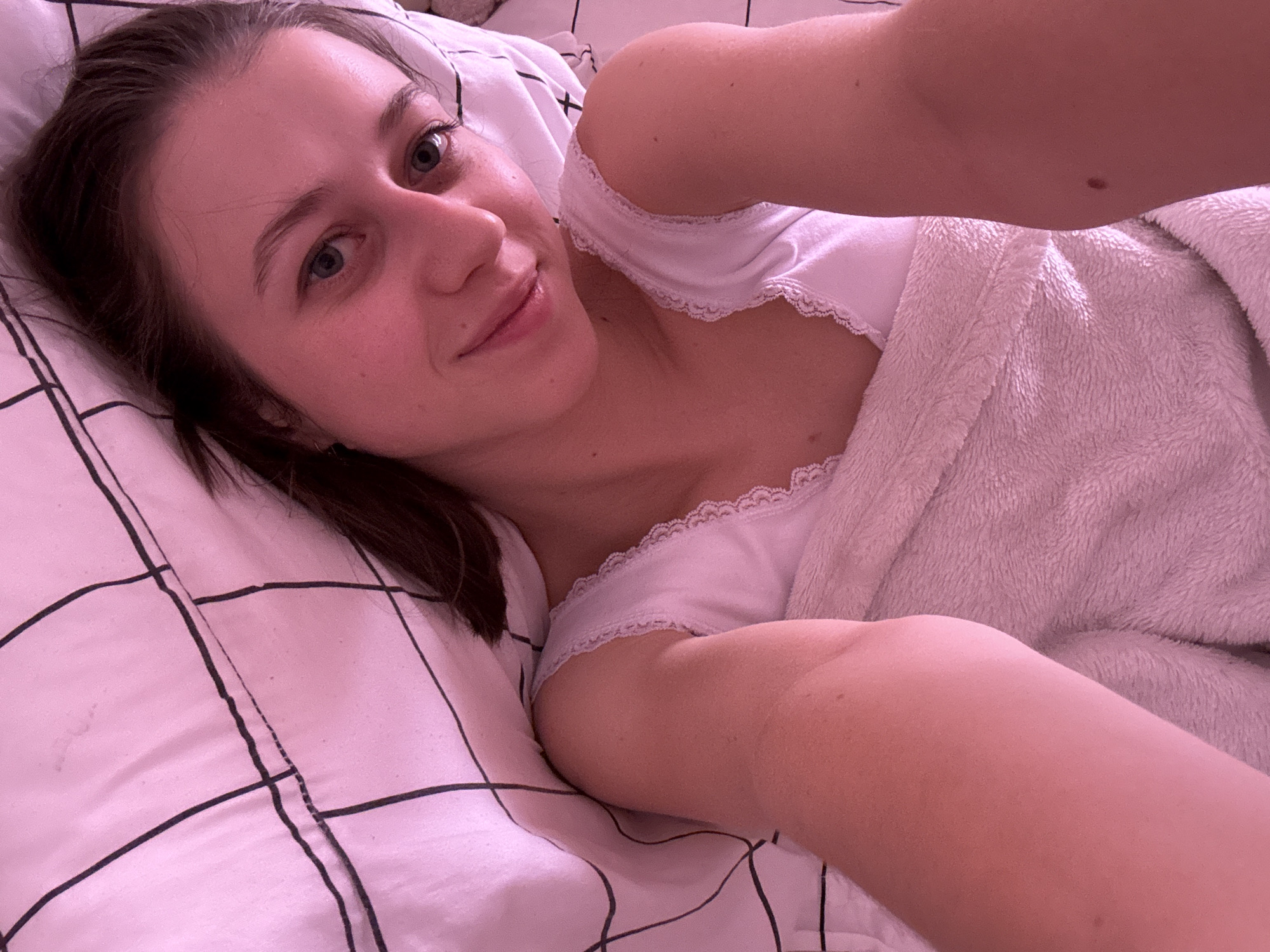 A young woman with dark brown hair, pulled back into a ponytail, is lying in bed, looking at the camera. She is wearing a white camisole with lace trim and is partially covered by a fluffy pink blanket. The bed linens are pink with a black grid pattern. The lighting is warm and pink-toned, suggesting it might be dawn or dusk, or artificial lighting. The overall scene depicts a private, intimate moment, possibly a selfie taken by the woman herself. There is no information to indicate the location in the city of Wörth am Main, Germany.