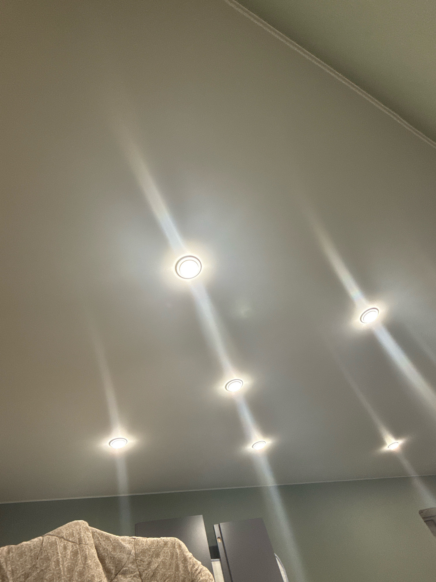 The image shows a ceiling with recessed lights and a portion of furniture or bedding in the lower left corner. There is no information in the image or provided context to suggest the location is Cheboksary, Russia. The scene is indoors, likely in a residential or office setting. The lighting suggests it may be evening or a dimly lit room. The ceiling appears to be a neutral color, possibly white or off-white. The furniture in the foreground is textured and has a patterned design.