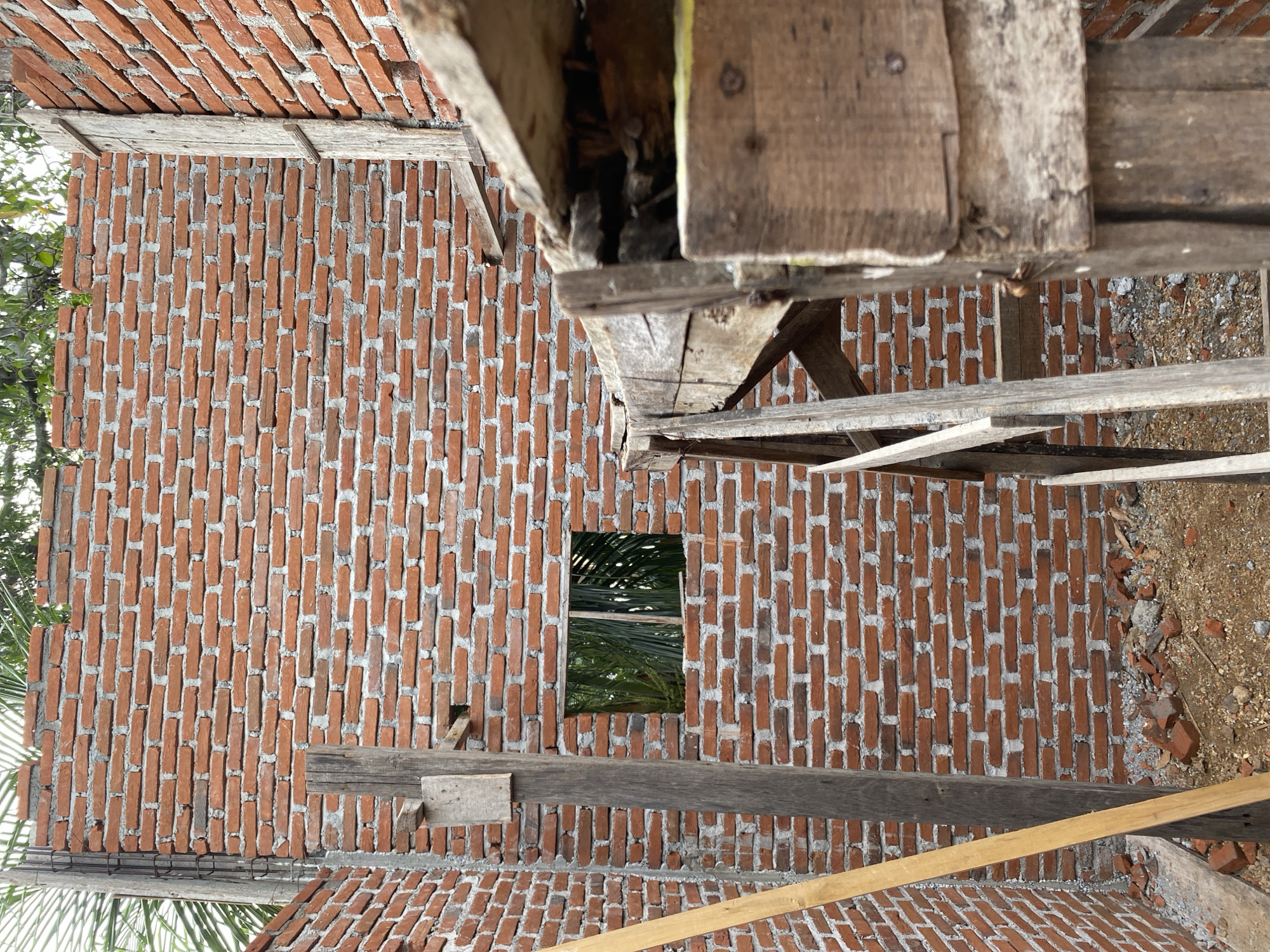 An exterior, eye-level, medium shot captures the construction of a brick wall. The wall is made of red bricks and is partially completed. A small, rectangular opening for a window is visible in the brick wall. Wooden scaffolding and supports are present around the wall. The ground is covered with dirt and debris, including broken bricks. Green palm leaves are visible through the window opening. The scene appears to be in an outdoor, possibly tropical, location.