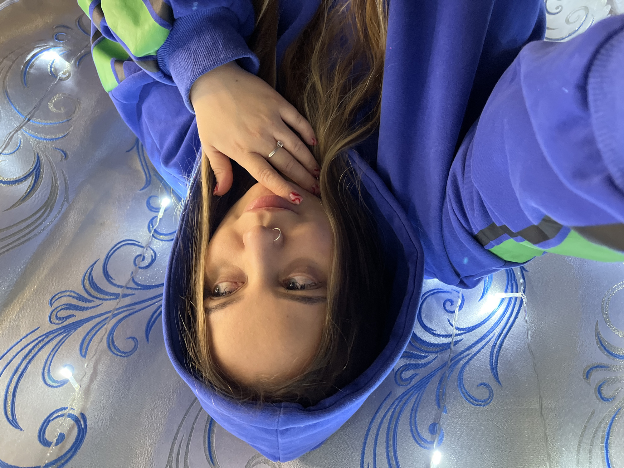 A woman with light brown hair and fair skin is lying down, looking directly at the camera. She is wearing a blue hoodie with green accents on the sleeves. Her left hand, adorned with a silver ring, is resting near her face. She has a silver hoop piercing in her left nostril. The background features a patterned surface of white and blue. There are also string lights visible, adding light to the scene. The location is unspecified and cannot be determined from the given image. The location is not likely to be in Novopokrovskaya, Russia.