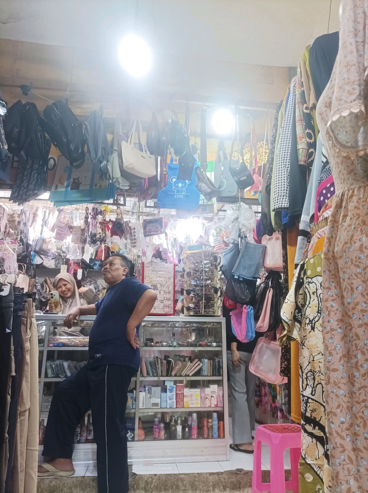 In Sumuragung, Indonesia, a man leans against a glass-enclosed display cabinet in what appears to be a small shop. He is wearing a dark blue t-shirt and black pants with a white stripe. His left hand is on his hip, and his right arm is resting on the cabinet. Behind him, a woman wearing a light brown hijab smiles and looks at a smartphone in her hand. She is standing behind the same cabinet. The shop is filled with merchandise. Bags of various styles and colors hang from the ceiling and hooks. Clothes are displayed on hangers and racks. The display cabinet contains shelves stocked with bottles, boxes, and other items, possibly toiletries or cosmetics. A bright light hangs from the ceiling, illuminating the shop. In the foreground, to the right, is a pink plastic stool.
