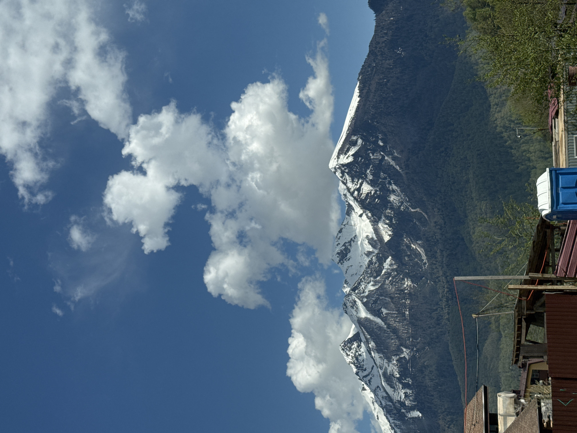 The image shows a snow-capped mountain range on the right side of the frame, with a forested area at its base. A section of the sky with clouds is visible on the left. In the foreground, on the right side, is a building with a red roof and a light blue object on top. The image appears to be taken in a location near a mountain range, with a building or structure in the foreground. The location is Unknown, Unknown.