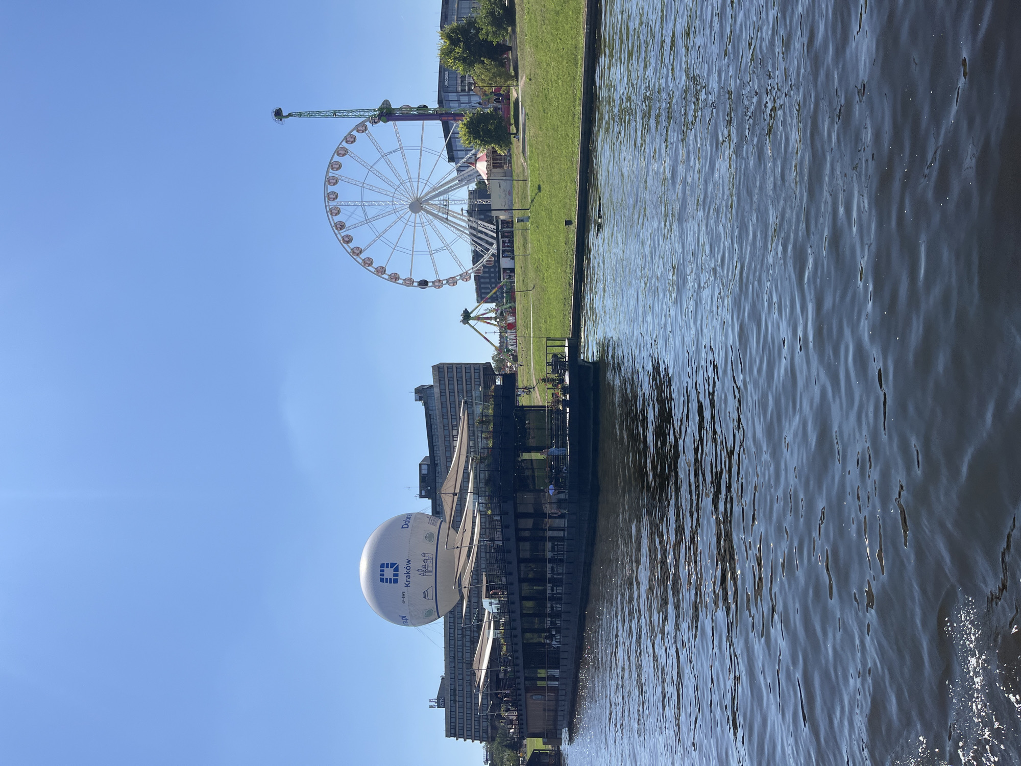 In Krakow, Poland, a modern Ferris wheel and a thrill ride stand prominently on a grassy embankment, overlooking a river. A large, inflatable white sphere, emblazoned with the text "Kraków," floats in the sky, attached to a building in the background. A long, dark boat with a covered upper deck and glass windows is moored on the river. People are visible on the boat's deck and on the riverbank. The water is choppy, reflecting the bright blue sky.