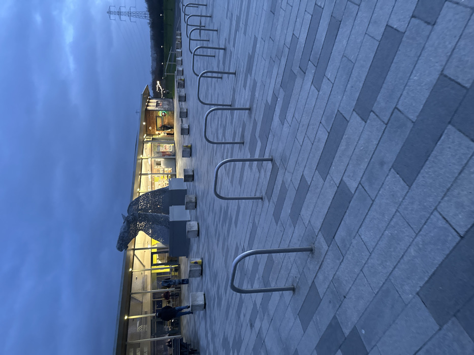 This image depicts an outdoor scene at The Helix, Falkirk, United Kingdom, at dusk or night, under an overcast, dark blue-grey sky.
In the foreground, a paved plaza composed of rectangular grey and darker grey pavers, arranged in a staggered pattern, extends towards the midground. Eight vacant, silver-colored, inverted U-shaped bicycle racks are uniformly spaced and aligned diagonally from the lower left to the middle right of the frame.
The midground features a large, illuminated metal sculpture of a horse's head, consistent with 'The Kelpies' artwork, positioned prominently. The sculpture exhibits an intricate, open mesh construction and is lit from within and below. Directly behind the sculpture is a single-story modern building, identified as a visitor center or retail/food outlet, characterized by extensive glass frontages and a flat roof with external lighting. The interior of the building is brightly illuminated, revealing details through the windows. A series of approximately eight uniform grey concrete bollards are arranged in a linear formation between the paved plaza and the building, with some topped by small yellow conical markers.
Multiple individuals are present: one person in dark clothing walks away from the camera towards the building's left entrance, another stands near a table to the far left of the building, and at least three additional figures are partially visible inside or near the building's entrances.
The background consists of a sloping grassy area behind the building, with a distant electrical transmission tower (pylon) visible against the sky on the far right. A dark treeline or dense vegetation forms the horizon line.