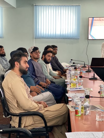 The image depicts a meeting or presentation in Shurandam, Afghanistan. Several men are seated around a long table, facing towards a projector screen displaying "Marketing Tools." They are all dressed in traditional Afghan clothing. There are drinks, including "Juicy Peach" juice boxes, water bottles, and glasses on the table. Microphones are also present. The room has blue-tinted blinds on a window. A laptop sits on the table near the presenter.