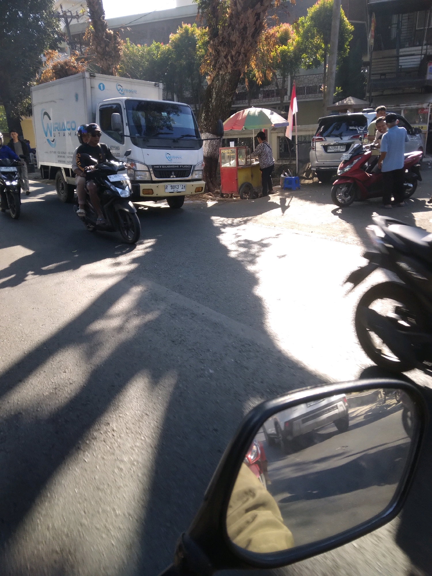 The image depicts a street scene in what appears to be a semi-urban or commercial area, possibly within Desa Pamijen, Kecamatan Sokaraja, Indonesia, judging by the context of the provided information. A white box truck with the logo "WIRIACO UTAMA" is parked on the right side of the road. Two individuals are on a black motorcycle in front of the truck, one driving and the other as a passenger. A red motorcycle is parked near a street food vendor's cart, which has a multi-colored umbrella and signage indicating "PENTOL THE SANG". A silver SUV is also parked in the vicinity. Several people are visible near the vendor and the parked motorcycles. Shadows of trees and other objects are cast on the asphalt road. The scene appears to be captured from the perspective of someone on a motorcycle, as indicated by the presence of a motorcycle mirror in the lower right portion of the frame, reflecting other vehicles on the road. The lighting suggests it is daytime, with sunlight casting distinct shadows.