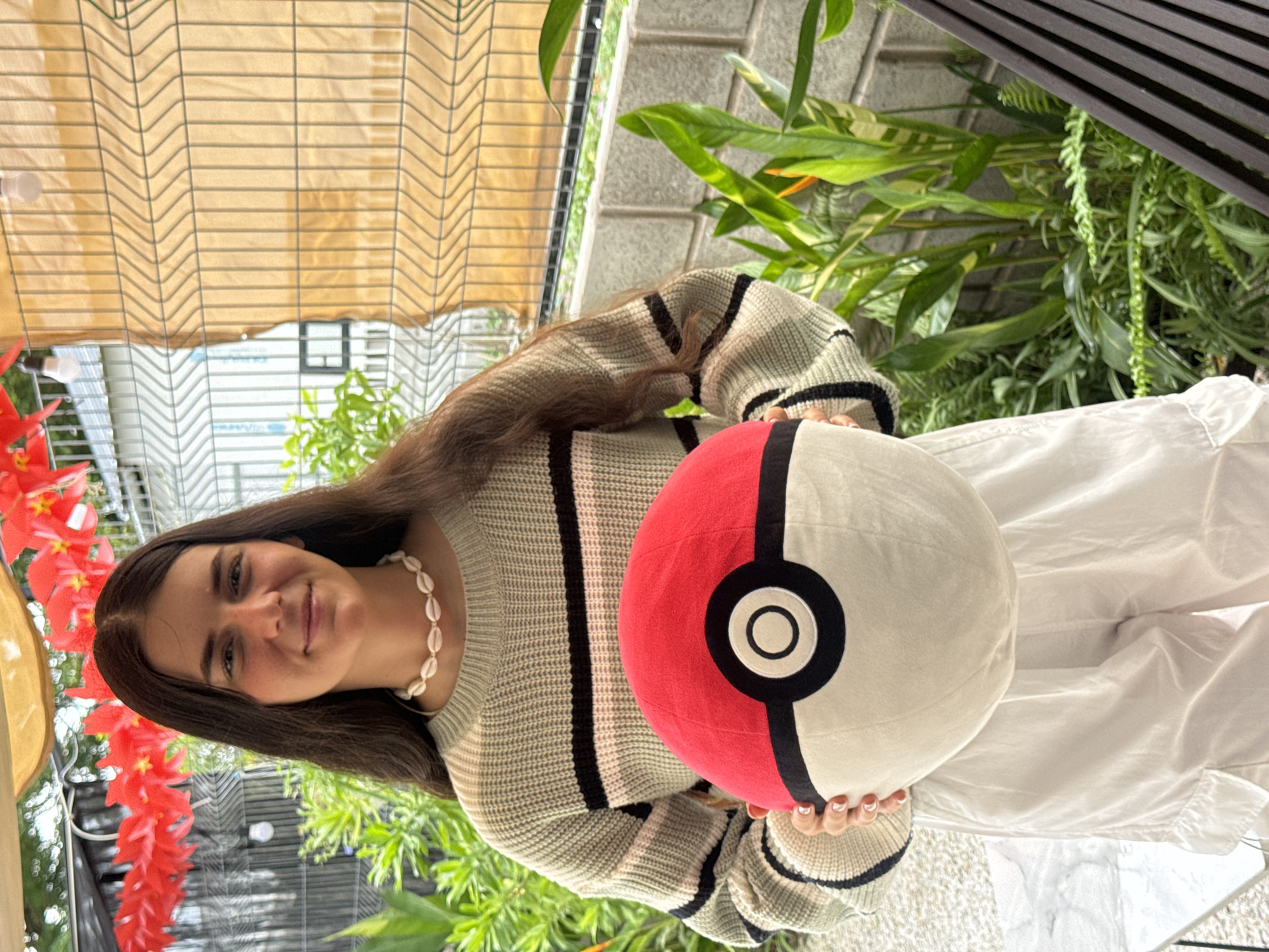 A young woman is holding a Pokemon ball pillow. The woman has long brown hair and is wearing a striped sweater and a seashell necklace. She is standing in what appears to be an outdoor setting with a variety of plants and flowers in the background. There is a structure with a tan woven covering overhead. The location is Phu Thuy Ward, Vietnam.