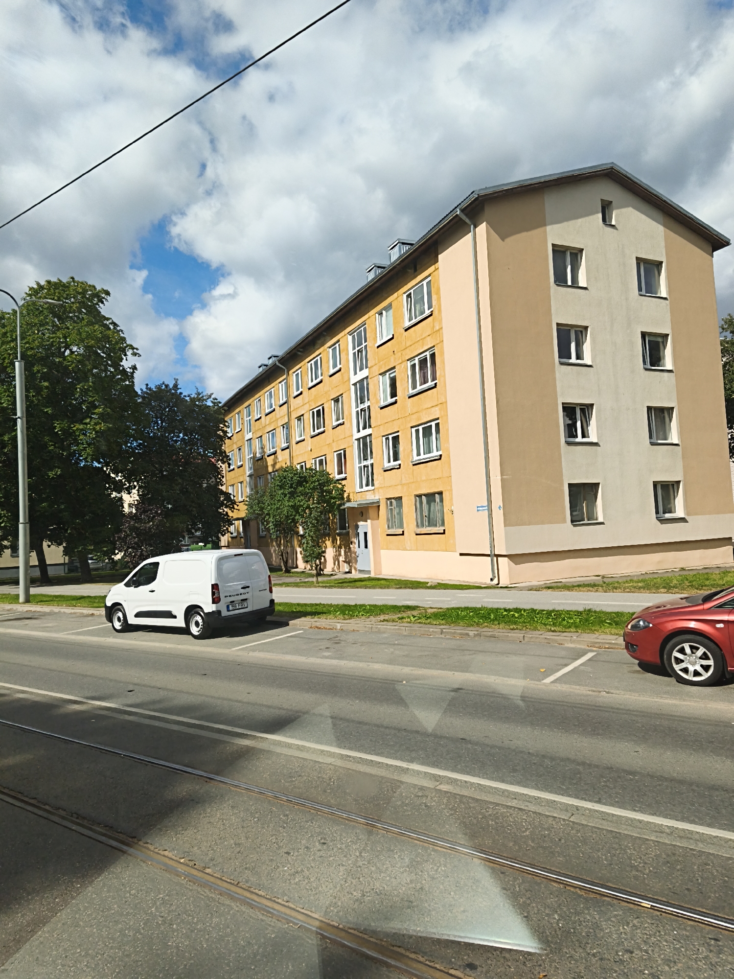 The image shows a long, multi-story apartment building with yellow and tan facades, seen from a street level perspective. The building has numerous windows, many with white frames, and a grey roof with dormer windows. To the left of the building, there are mature trees. In the foreground, a street with tram tracks runs horizontally across the lower part of the image. Parked on the side of the street in front of the building is a white van, a Peugeot model, with license plate "155 ERV". A red car is partially visible on the right side of the frame, also parked on the street. The sky is partly cloudy with patches of blue. A power line or wire is visible stretching across the upper portion of the frame. The environment suggests an urban residential area in Tallinn, Estonia, likely a slightly overcast day.