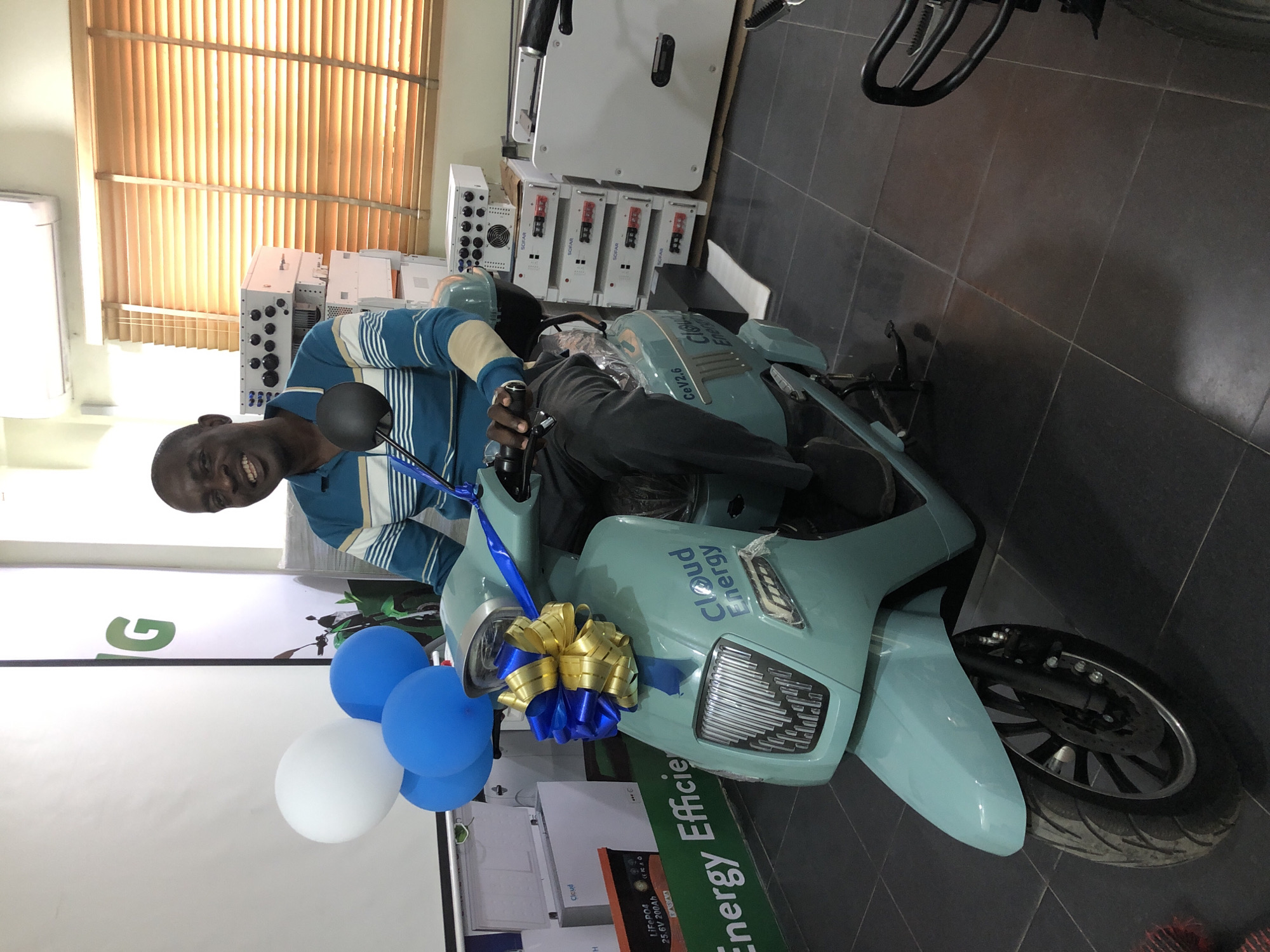 In a well-lit indoor space, possibly a showroom in Ikeja, Nigeria, a Black man in his 40s or 50s is seated on a light teal electric scooter adorned with a large golden and blue bow. He wears a blue and white striped long-sleeved shirt and dark pants. The scooter has the "Cloud Energy" logo visible on its front. In the background, stacked white boxes, some with "SOFAR" branding, and what appear to be battery units are arranged, suggesting a display related to energy solutions. A large white banner with a green logo and text is partially visible to the left, and the room has blinds covering a window on the right. Three balloons, one white and two blue, are tied to the scooter's handlebars. The floor is tiled in dark grey.