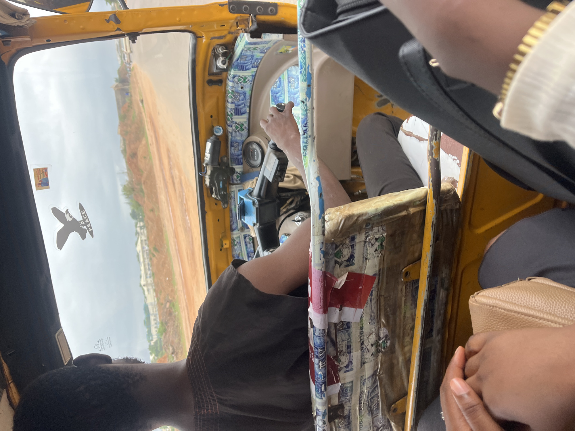 The image provides an interior view of a vibrant yellow vehicle, likely a tricycle taxi or "Keke Napep," from a passenger's perspective, looking towards the driver and the road ahead. The scene is set in Galadimawa Village, Nigeria, during the daytime under an overcast sky.
In the foreground to the right, a passenger's hands and arm are visible, showing light brown skin, a light-colored long-sleeved top, and a gold bracelet. They are holding a light brown, textured handbag. On the left, another passenger's shoulder and the back of their head are seen, wearing a dark brown or black shirt with subtle reddish stripes.
The driver, a person with dark skin and short black hair, is positioned at the front, with their arm extended to grip the vehicle's handlebar-style steering mechanism. The dashboard and interior panels are intricately decorated with blue and white patterned material, possibly fabric or paper, and sections of red tape. A speedometer is partially visible on the dashboard.
Through the front windshield, a wide dirt road with reddish-brown earth is visible, extending into the distance. Sparse green vegetation lines the road, and several light-colored buildings can be seen further away under a bright, cloudy sky. Two stickers are affixed to the windshield: a prominent black silhouette resembling a bird or airplane with the word "PEACE" below it, and a smaller rectangular sticker with unreadable text. The yellow frame of the vehicle shows some wear and tear.