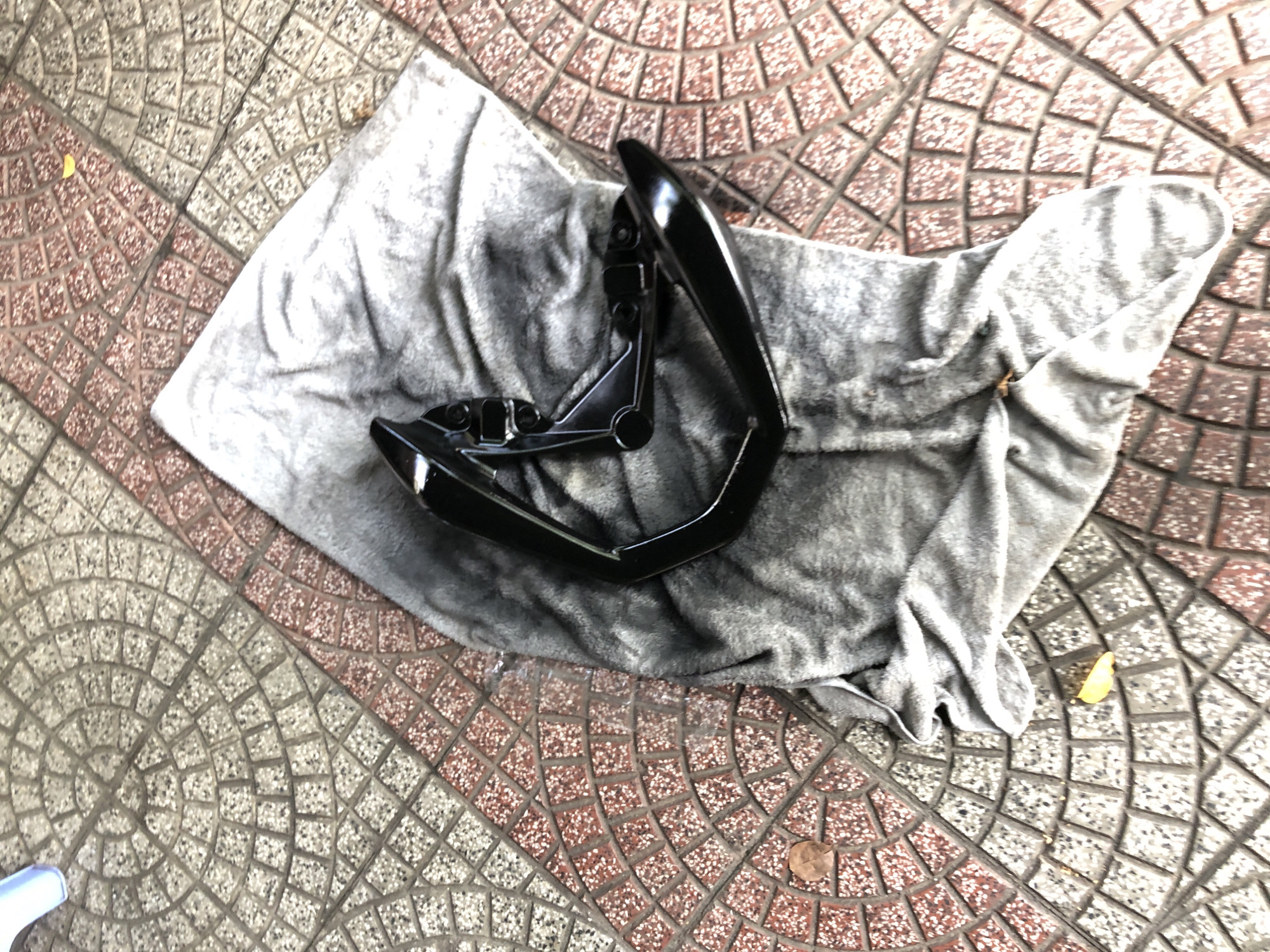A close-up, top-down shot captures a pair of black motorcycle footpegs resting on a grey cloth. The cloth is laid out on a paved surface composed of interlocking, circular patterned tiles. These tiles are primarily a light grey with concentric circles and a contrasting reddish-brown border. A portion of a blue object is visible in the upper left corner, outside the main focus. The scene is set on a sidewalk or pedestrian area in Đà Nẵng, Vietnam.