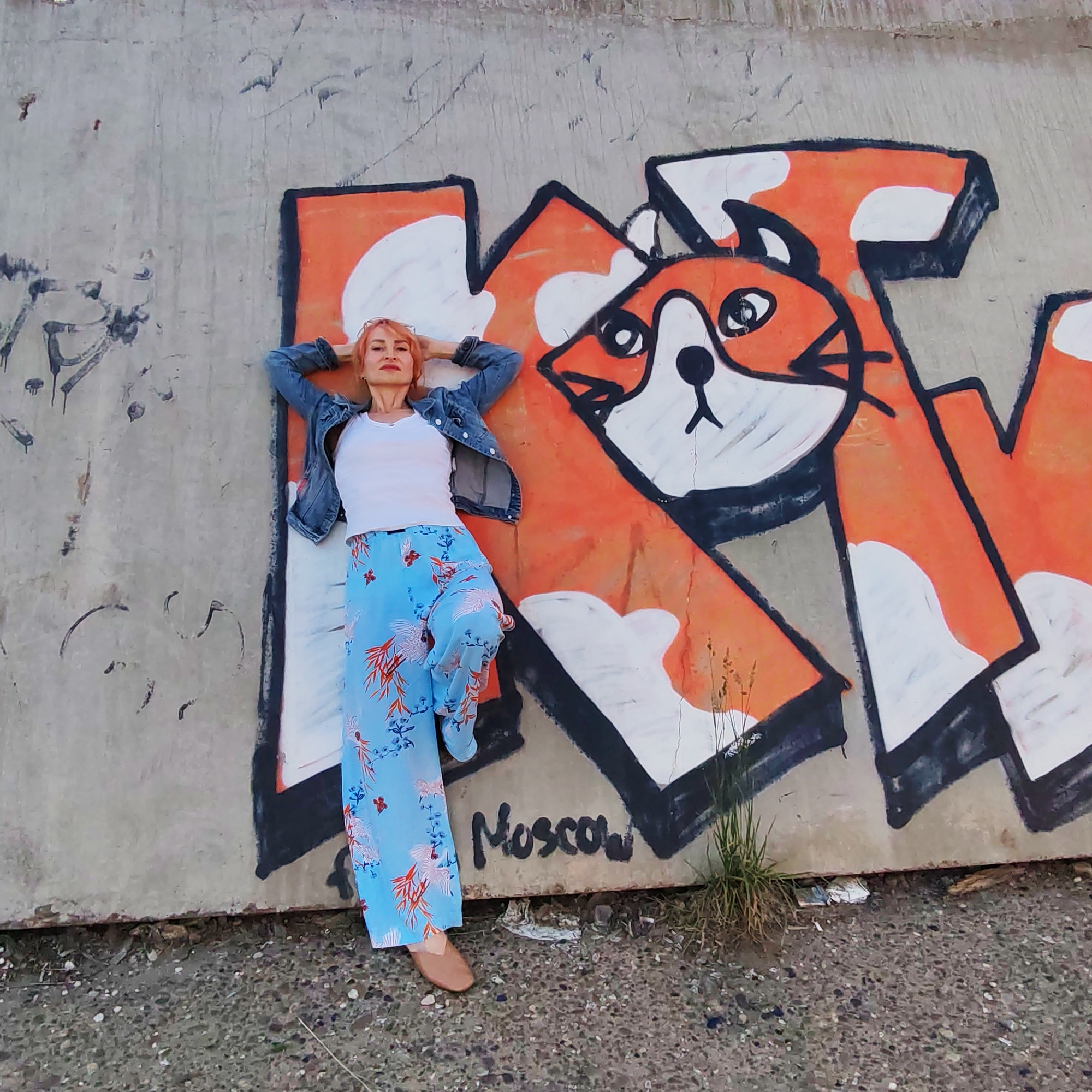 A woman with short, reddish-blonde hair is lying on her back against a concrete wall featuring graffiti. She is wearing a denim jacket over a white tank top and loose, blue, floral-patterned pants. Her hands are behind her head, and her legs are bent. The graffiti on the wall depicts a stylized fox's face within orange and white shapes, with the word "Moscow" written below it in black. The scene appears to be outdoors in an urban environment, with gravel and some sparse vegetation on the ground. The concrete wall shows signs of age and other, less defined graffiti. The image does not provide specific geographical information to pinpoint a location within Chaikovsky, Russia.