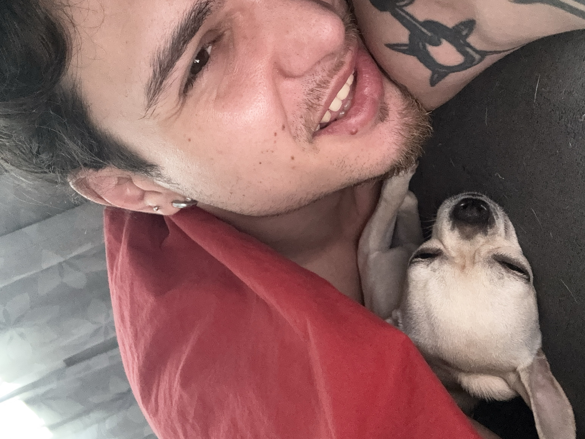 The image is a selfie taken from a close-up, slightly upside-down perspective. A man with dark hair, brown eyes, and stubble is visible in the upper left corner, smiling slightly. He has a silver hoop earring in his left ear and a tattoo on his right shoulder. Cradled against his neck is a small, light-colored dog, possibly a Chihuahua, with its eyes closed, appearing to be sleeping. A red cloth or blanket is draped across the lower left of the frame. The background is indistinct but suggests a casual indoor setting. There is no indication of being located in Sofia, Bulgaria.