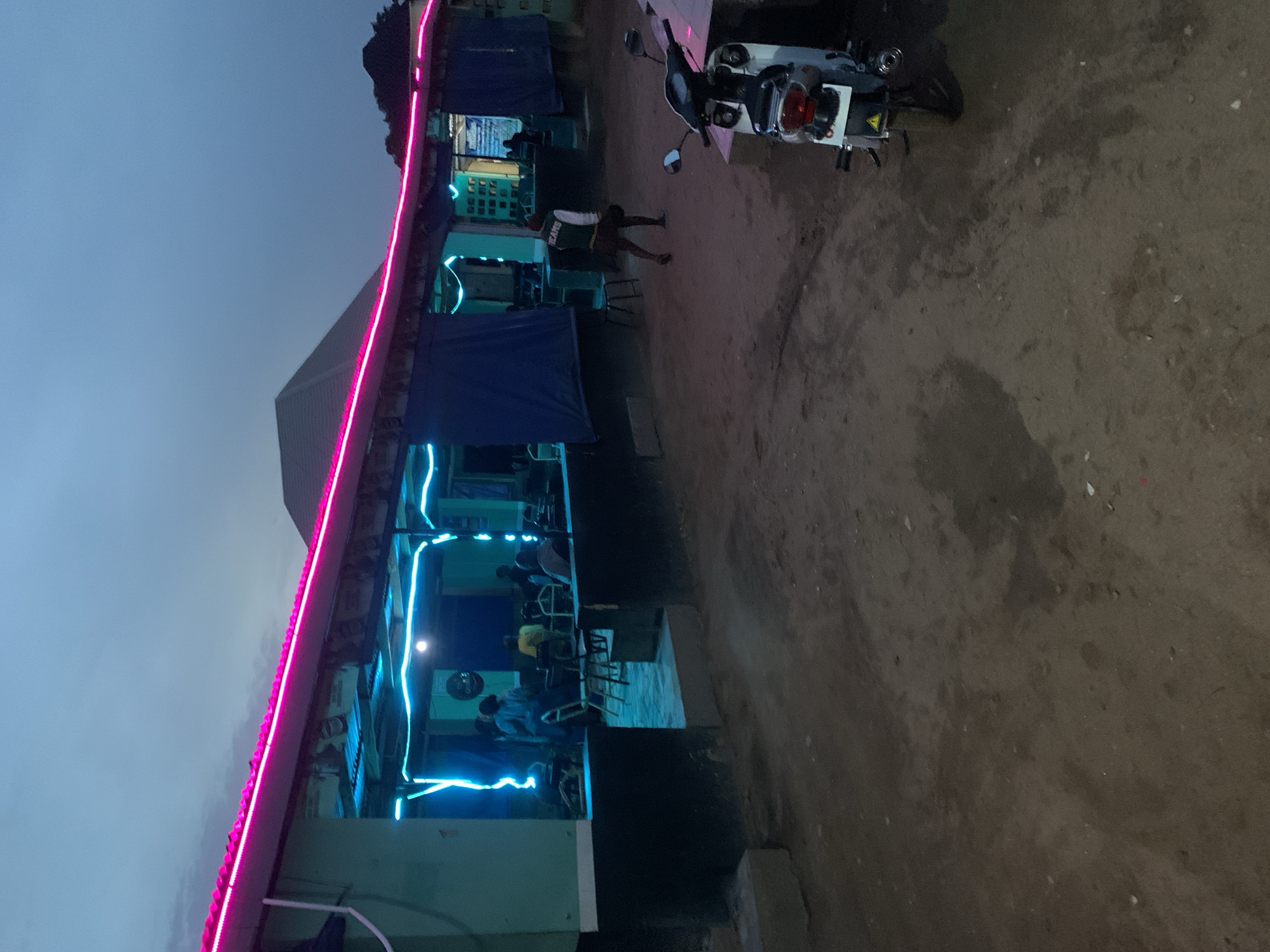 An outdoor scene at night in New Stadium, Nigeria, depicts a building with a pink-lit roofline and blue neon lights accentuating its architectural features. Inside the building, people are seated around tables. Outside, a parked motorcycle is visible, along with a person in a white and green jacket. The ground is dirt and gravel. The sky is dark.