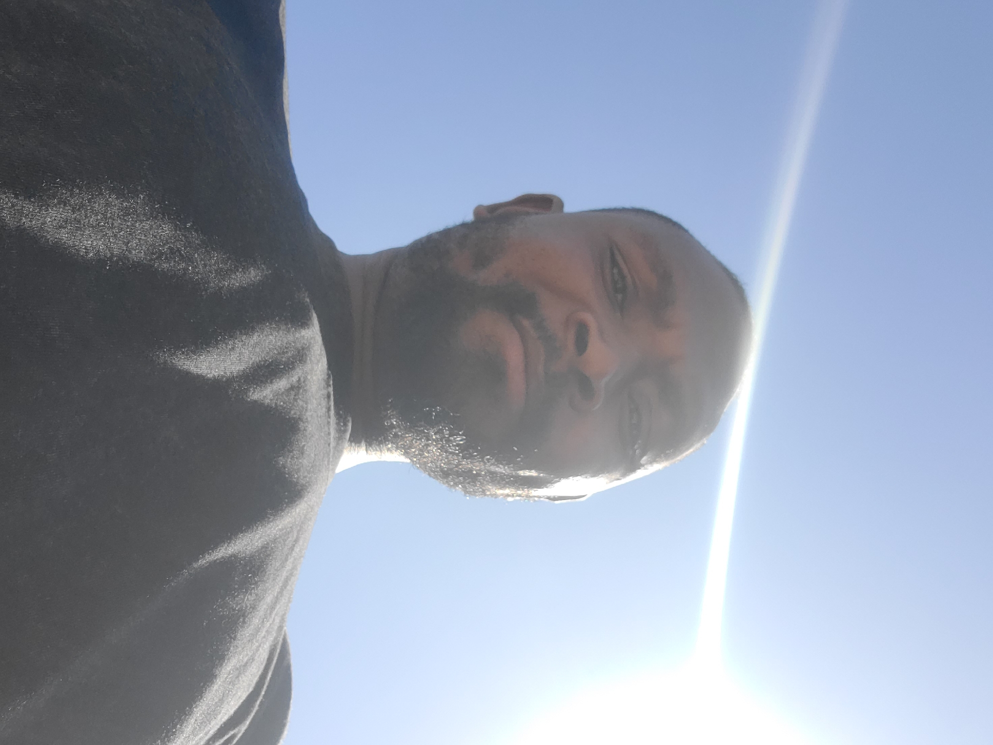 A low-angle, medium shot captures a Black male from the shoulders up, against a bright blue sky with a lens flare from the upper right. He has a closely cropped haircut and a short beard. He is wearing a dark gray crew-neck t-shirt. The perspective suggests the photo was taken from below, possibly by him. The scene appears to be outdoors in a clear, sunny environment. The location in Gura Top, Nigeria is not evident from the image itself.