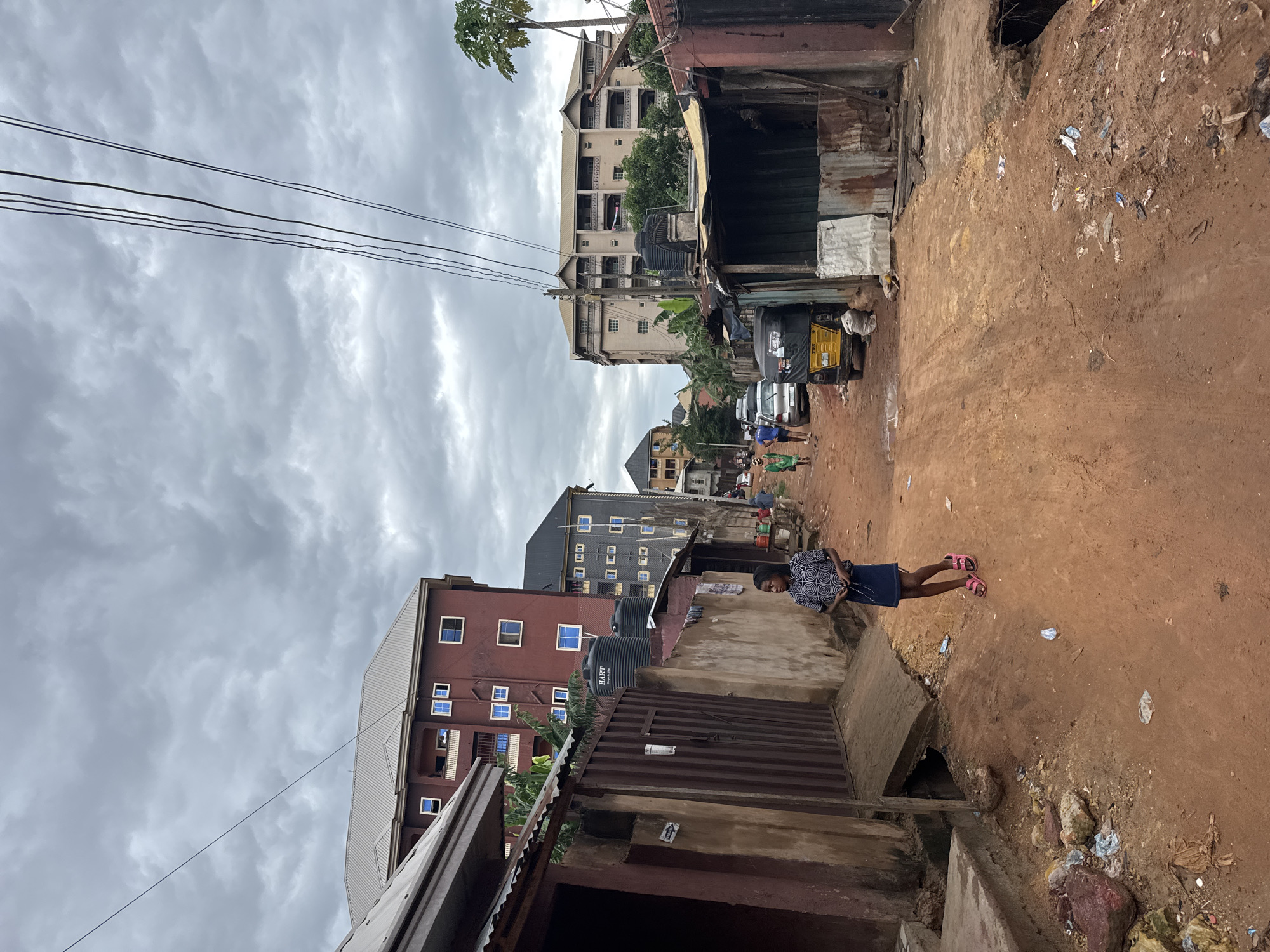 The image depicts a street in Nkpor, Nigeria, under a cloudy sky. The street is unpaved with reddish-brown dirt and scattered litter. A young girl stands near a concrete drain, wearing a patterned top, dark skirt, and pink sandals. Buildings of varying heights line the street, including multi-story structures with exposed water tanks and corrugated metal shacks. Power lines stretch across the sky. Further down the street, a tricycle and other vehicles are parked, and a few people are visible.