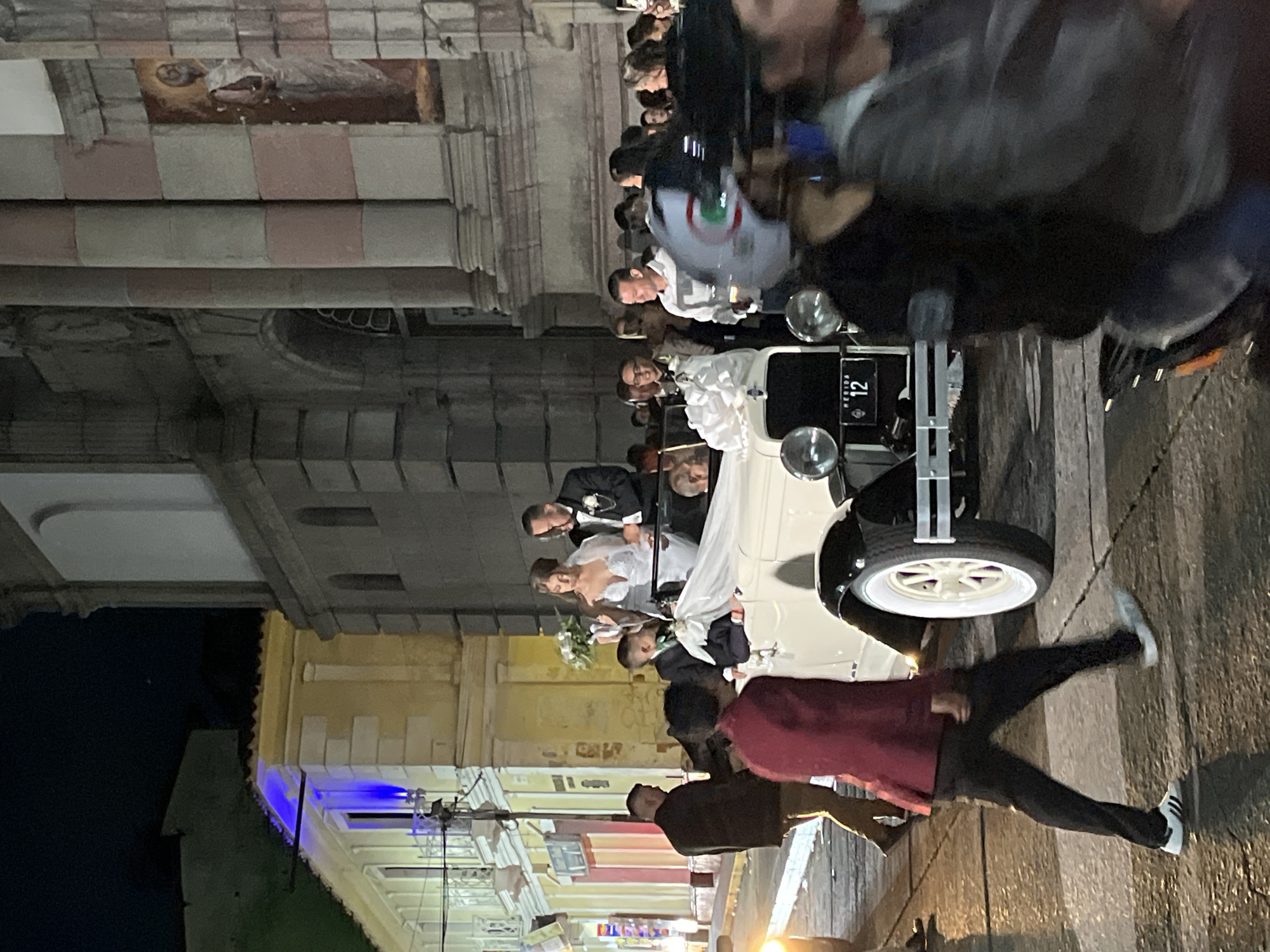 This is a nighttime, eye-level, medium shot photograph capturing a wedding scene on a city street in Mérida, Venezuela. In the foreground, several people are visible. A person in a maroon jacket and black pants walks past a classic white car. The car is decorated with white fabric and a bouquet. A bride in a white wedding dress and a groom in a black tuxedo are standing in the car, appearing to be the focus of the event. Other wedding guests and onlookers are gathered around, some partially obscured. In the background, a large building, likely a church or historical structure, is lit, revealing architectural details such as columns and windows. Adjacent to this building is a yellow-toned building with blue lighting, suggesting a commercial establishment or residential area. The street appears to be paved, and streetlights are faintly visible in the distance. The overall atmosphere is celebratory, with a sense of movement and activity as people interact with the wedding party and the car.