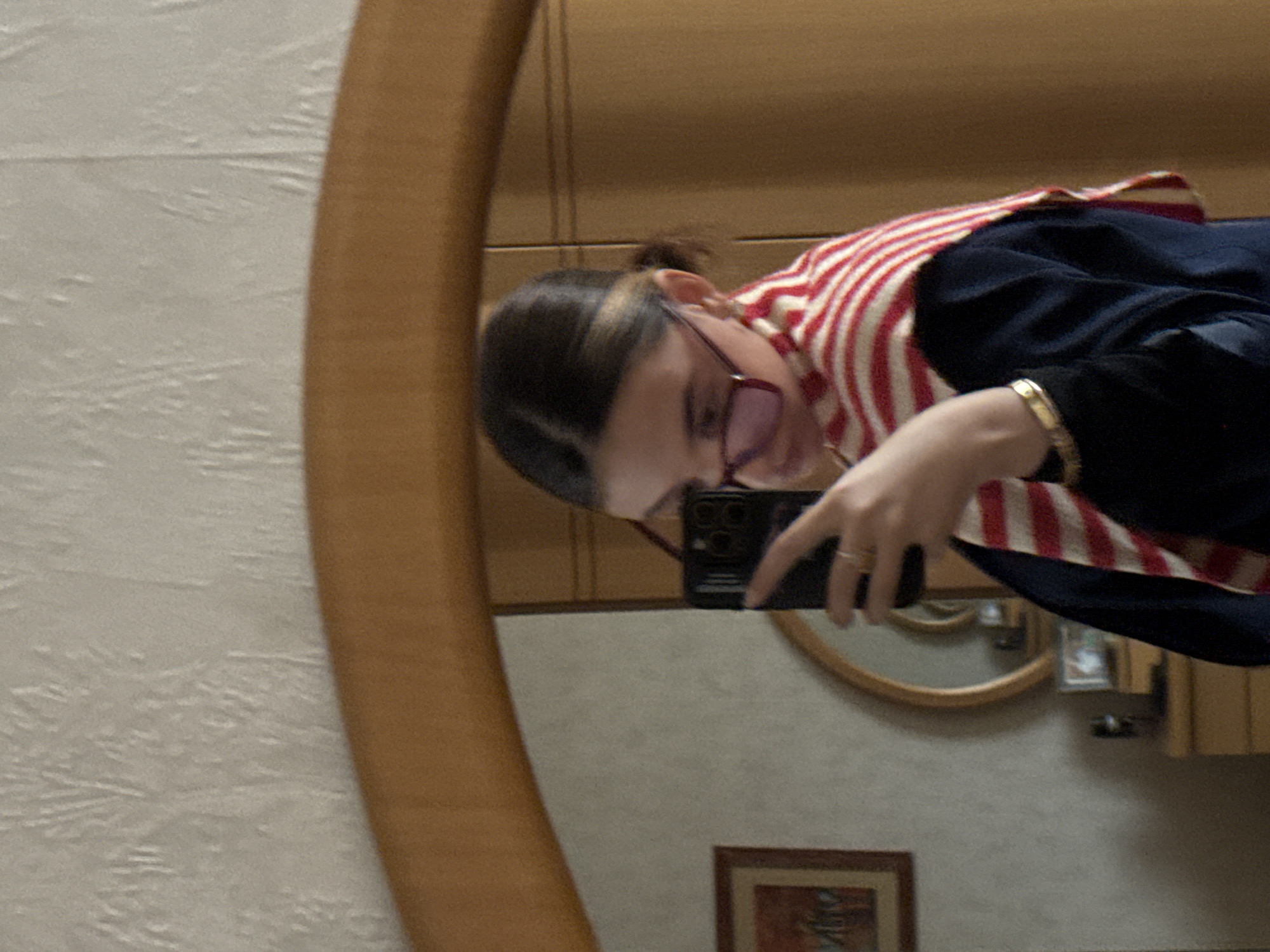 An individual is shown taking a mirror selfie in an indoor residential setting in Yerevan, Armenia. The subject, a young adult with dark hair tied back and wearing red-framed glasses, holds a black smartphone in their right hand, angled to capture their reflection. They are dressed in a dark top or jacket and a red and white striped scarf. A gold-colored bangle is visible on their left wrist, and a ring on the right ring finger. The large, arched mirror, framed in light wood, reflects a room with light-colored, textured wallpaper. Visible within the reflection are a framed picture with a brown frame displaying an abstract image on the left, and a partial view of a light brown wooden dresser or cabinet below it. A smaller, circular mirror with a light wooden frame is also reflected on the dresser. To the right, a section of a wooden wardrobe or closet is present.