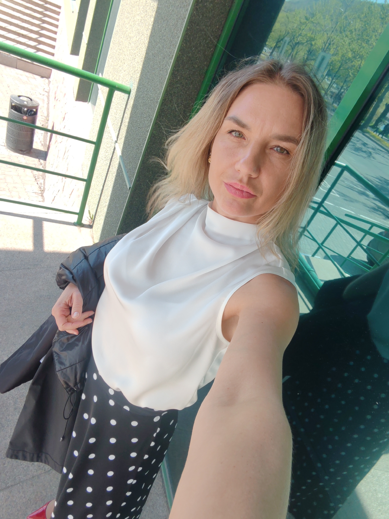 A blonde woman with light skin and light green eyes stands on a balcony. She is wearing a white sleeveless top and a black skirt with white polka dots. She is holding a black jacket over her left arm. Her right arm is extended towards the camera. In the background, there is a green railing, a grey wall, and a reflection in a green-tinted window. To the left of the woman, there is a grey trash can and a staircase. The scene is outdoors during the day.