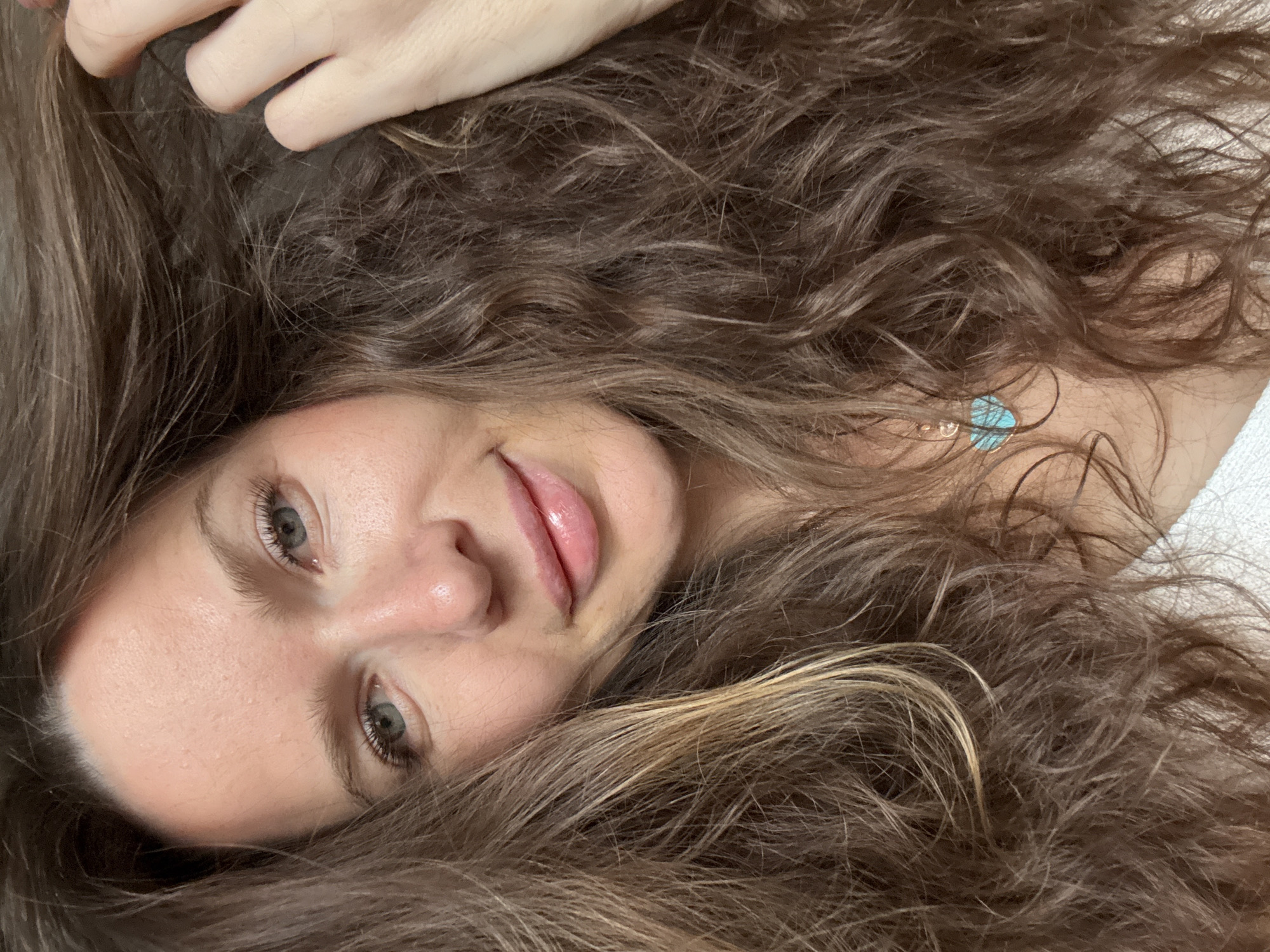 The image is a close-up, high-angle portrait of a young woman lying down, her head rotated clockwise, surrounded by her voluminous, long, wavy brown hair. Her left hand is visible near the top of the frame, fingers gently touching her hair. She has light-colored eyes, possibly green or hazel, with long eyelashes. Her lips are slightly parted in a subtle, gentle smile or smirk, coated with a glossy finish. Her skin appears clear. Nestled among her hair on her chest, a small, light blue pendant or charm on a delicate chain is visible. The background suggests a soft, light-colored surface, possibly bedding or a cushion, indicating an indoor setting. The lighting is soft and even, suggesting either natural indoor light or diffused artificial light, but the specific time of day is not evident. Her expression conveys a sense of relaxation, ease, and subtle playfulness. No text is visible in the image. The location context for this media is Moscow, Russia.