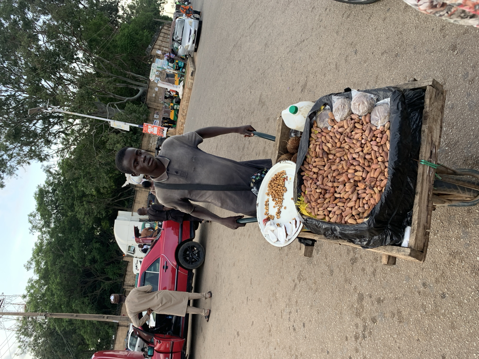 A man stands behind a cart loaded with goods, holding a long pole. The cart is filled with a large quantity of dried dates, some of which are packaged in plastic bags. A white plate on the cart holds smaller quantities of nuts and what appear to be slices of coconut. A white plastic jug with a green lid is also visible in the cart. The man wears a dark grey t-shirt. The scene is a street in Jos, Nigeria, with other people and vehicles present. A red car is parked to the left of the cart, and another person in a beige outfit is seen leaning into the car. The background shows a street lined with trees and buildings, with several tuk-tuks and cars suggesting a busy urban environment. The lighting suggests it is daytime, possibly late afternoon or early evening. There is no visible text in the image that is clearly legible.