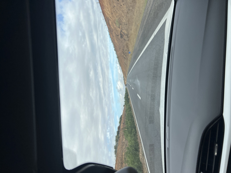The image displays a straight asphalt highway, observed from the interior of a vehicle through its windshield. The top of the vehicle's dark headliner and the lower portion of its white dashboard, including air vents, are visible in the foreground. The road has two lanes marked by white dashed lines for the median and solid white lines defining the shoulders. Isolated patches of moisture are present on the road surface. On either side, reddish-brown earthen slopes or embankments rise, covered with sparse green and dry vegetation. A blue road marker sign is visible on the right shoulder. The sky is predominantly overcast with white and gray clouds, revealing intermittent patches of blue sky. Distant vehicles are discernible on the road ahead. This scene is located in Ait Ouallal, Morocco.