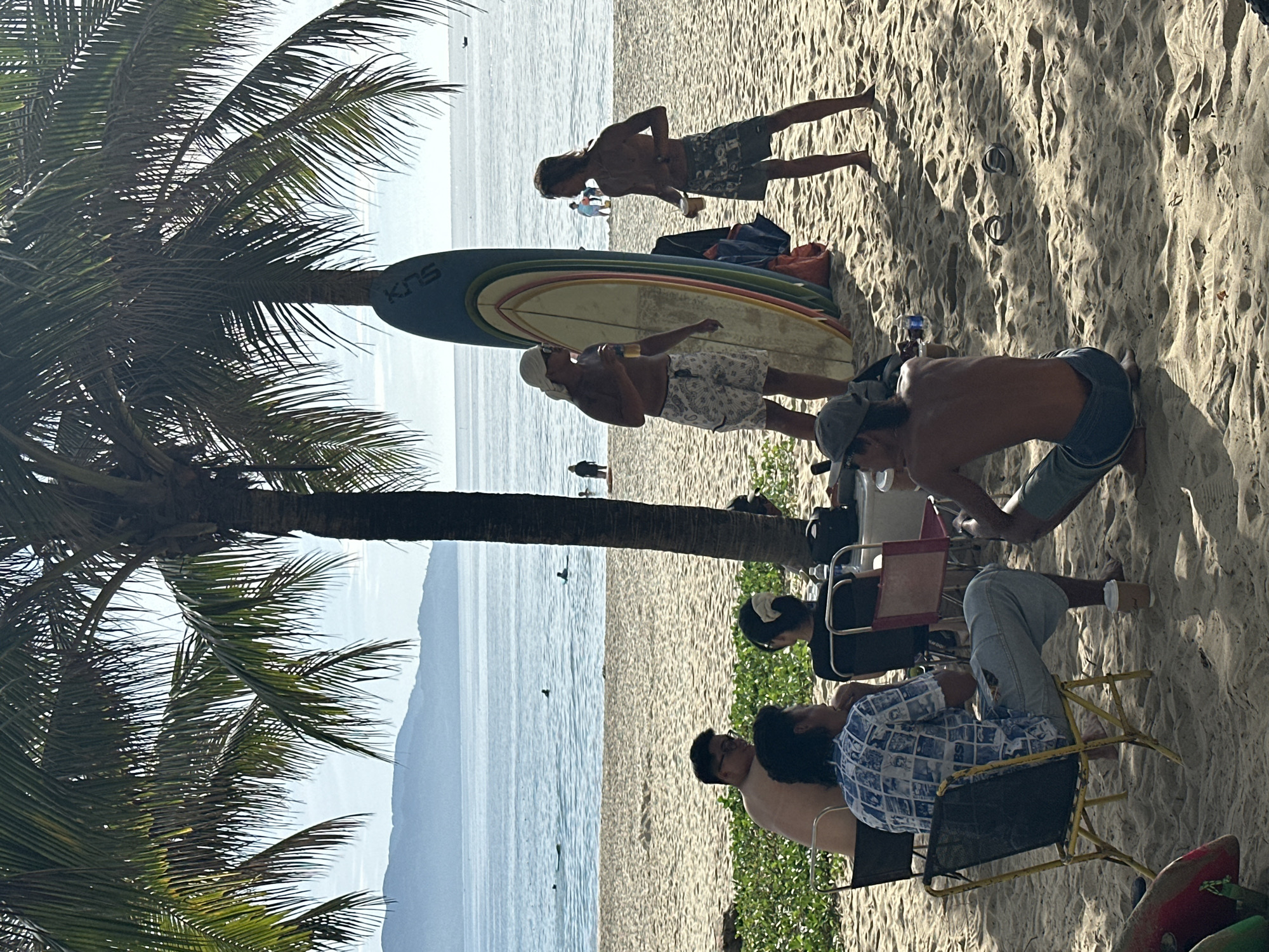 This image depicts a beach scene in Da Nang, Vietnam, likely My Khe Beach, given the common presence of palm trees and surfboards. Several people are gathered on the sand under the shade of palm trees, with the ocean and distant hills visible in the background. In the foreground, four individuals are seated on chairs. One man in a blue and white patterned shirt and light blue pants is sitting in a yellow chair facing away from the camera. To his left, another man is seated, mostly obscured, wearing glasses. A woman with dark hair tied back with a light-colored scrunchie is seated next to the man in the patterned shirt. In the mid-ground, two shirtless men are standing and talking. One is wearing a white bandana and patterned shorts, holding a drink, and standing next to a colorful surfboard propped against a palm tree. The other man is wearing patterned shorts and also holding a drink. Further back, several people are visible near the water, some appearing to be surfing or wading. The sand is light and textured, with some sparse green vegetation in the lower left corner. The overall atmosphere suggests a relaxed, casual day at the beach.