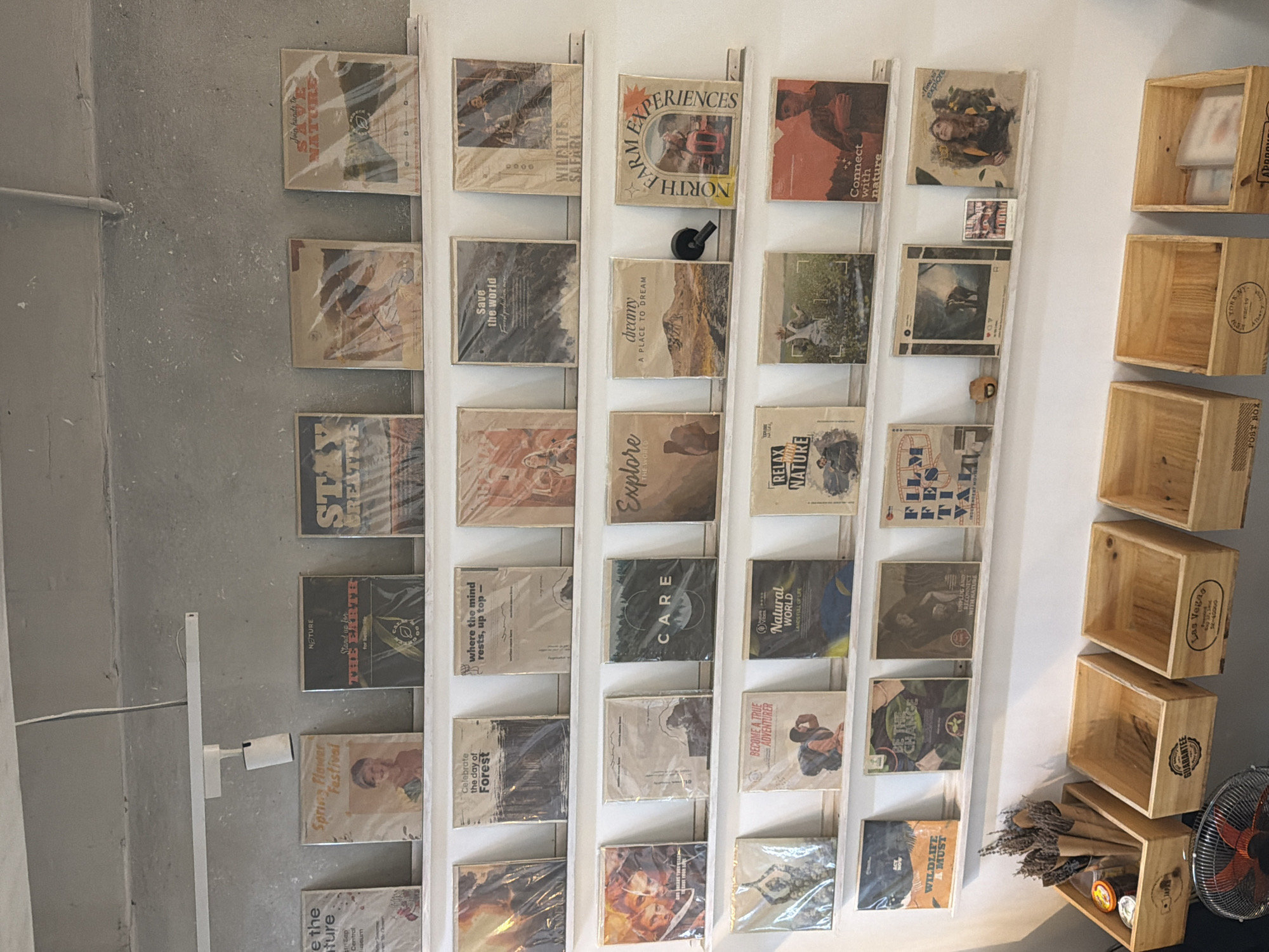 The image displays a wall of shelves filled with framed prints or artwork, predominantly featuring nature and travel themes. The items are arranged in neat rows, with each piece individually wrapped in plastic. The color palette of the prints leans towards earthy tones and vintage aesthetics. Below the shelves, several wooden crates are visible, some empty and others containing small items like dried flowers, jars, and what appear to be more of the framed prints. A black fan is positioned in the lower left corner of the image. The overall impression is that of a retail or display space, possibly a shop or gallery. The lighting suggests an indoor environment, with a white fixture attached to the ceiling. The backdrop is a plain, light-colored wall. The location is identified as Nam Nha Trang Ward, Vietnam.