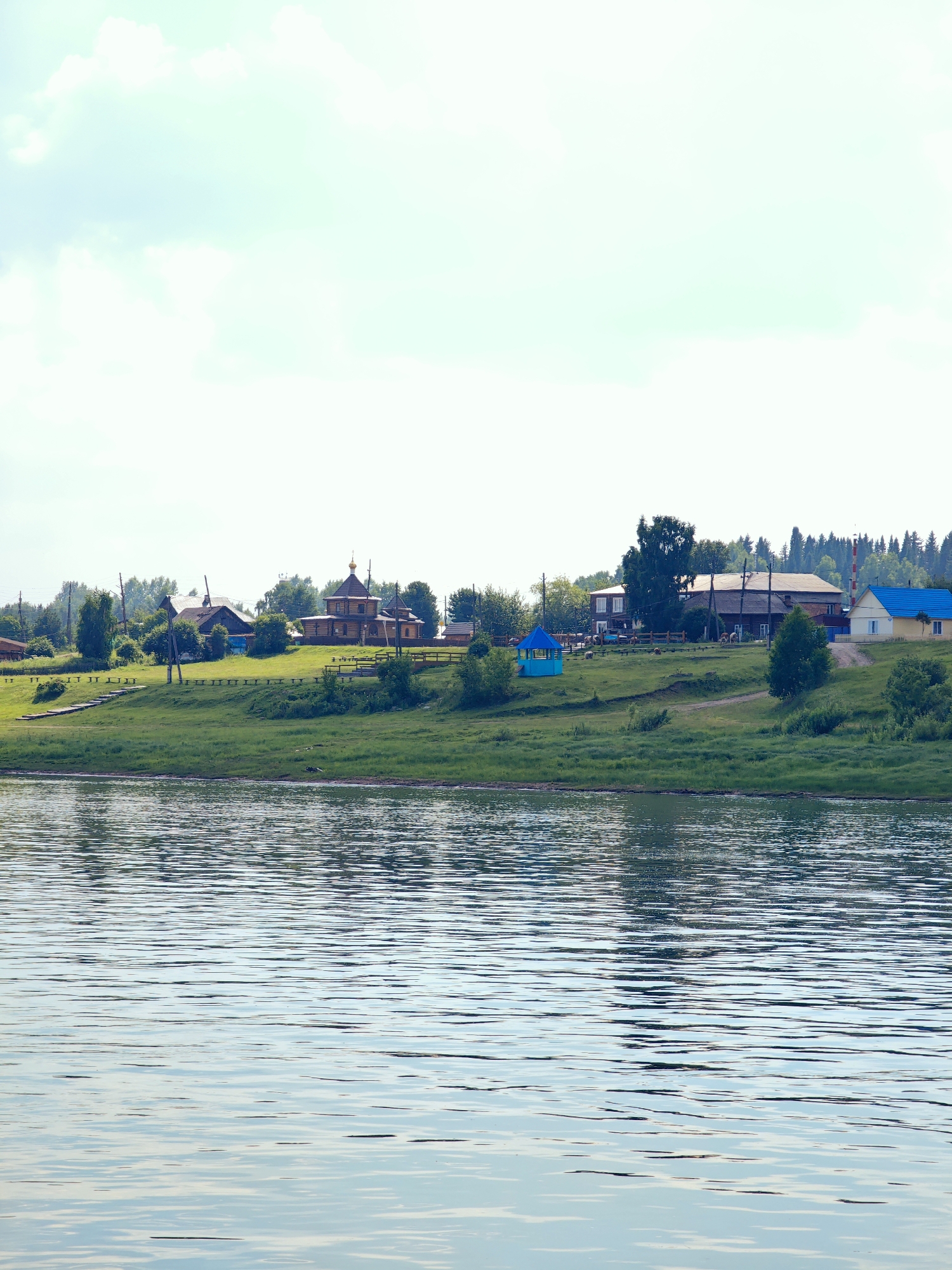 The image depicts a tranquil scene of a riverside village in Ust-Kem, Russia. In the foreground, a body of water with gentle ripples reflects the sky and the distant landscape. The middle ground features a grassy embankment leading up to a cluster of buildings. Among these structures, a prominent wooden church with a dark, tiered roof and a golden dome stands out. Other buildings, including houses and a bright blue gazebo, are visible further along the embankment. The background is filled with lush green trees and a hazy sky with scattered clouds. The overall impression is one of a peaceful rural settlement on a clear day.