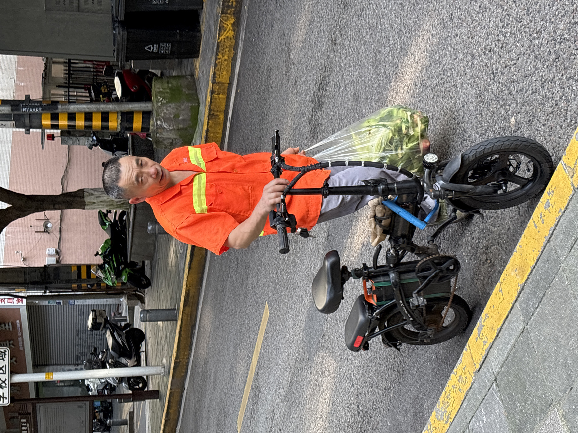 A middle-aged Asian man wearing a bright orange work shirt with reflective yellow stripes and light gray pants walks his black electric bicycle down a city street. The man is holding the handlebars with his right hand and carrying a large clear plastic bag filled with green vegetables in his left hand. The bicycle has a black seat and a secondary seat or storage area behind it. The man's left foot is on the pedal. The scene is an asphalt street with a yellow painted curb line. In the background, there are parked motorcycles, a green sport bike, trash cans, and building facades. The image appears to have been taken in Yuzhong District, China.