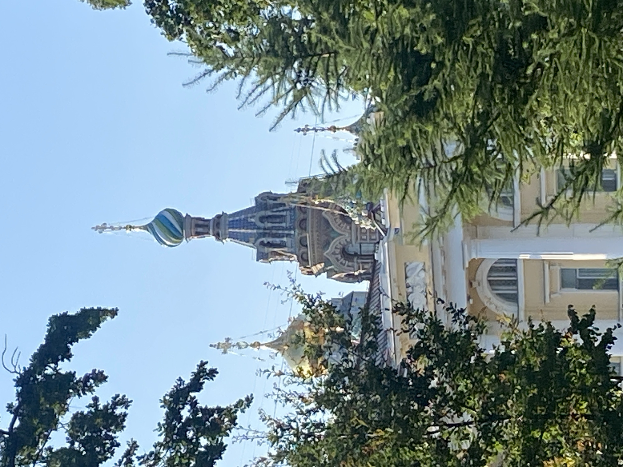 The image shows a section of the Church of the Savior on Spilled Blood in Saint Petersburg, Russia. The foreground is dominated by lush green trees, with branches partially obscuring the view of the church. Behind the trees, the golden dome of one of the church's smaller chapels is visible, topped with a cross. To its right, a larger, more elaborate dome with alternating green and blue stripes, also topped with a cross, rises prominently against a clear blue sky. The church's intricate architecture, with its colorful mosaics and detailed ornamentation, is partially visible. The building's facade, in a pale yellow color, features classical columns and arched windows. The overall scene depicts a sunny day with a clear sky, showcasing a part of this iconic religious landmark amidst natural foliage.