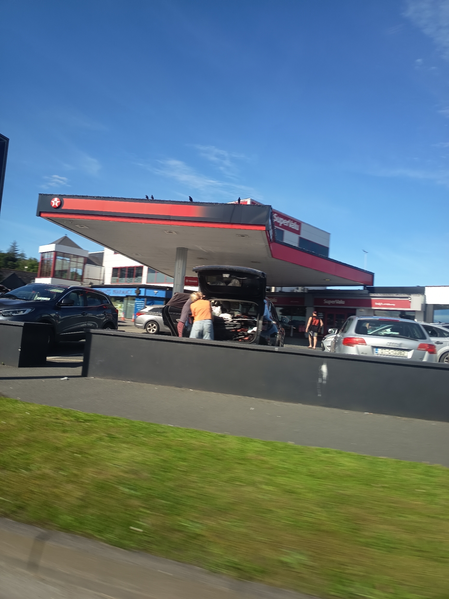 The image shows a busy scene at a Supervalu-branded gas station in Killarney, Ireland. The gas station has a distinctive red and white overhang with a prominent Supervalu logo. Several cars are present, including a dark SUV with its trunk open, and a silver hatchback. People are seen interacting with the cars, possibly loading or unloading items. The sky is clear and blue, indicating a sunny day. The overall environment suggests a typical retail and refueling area within the town.