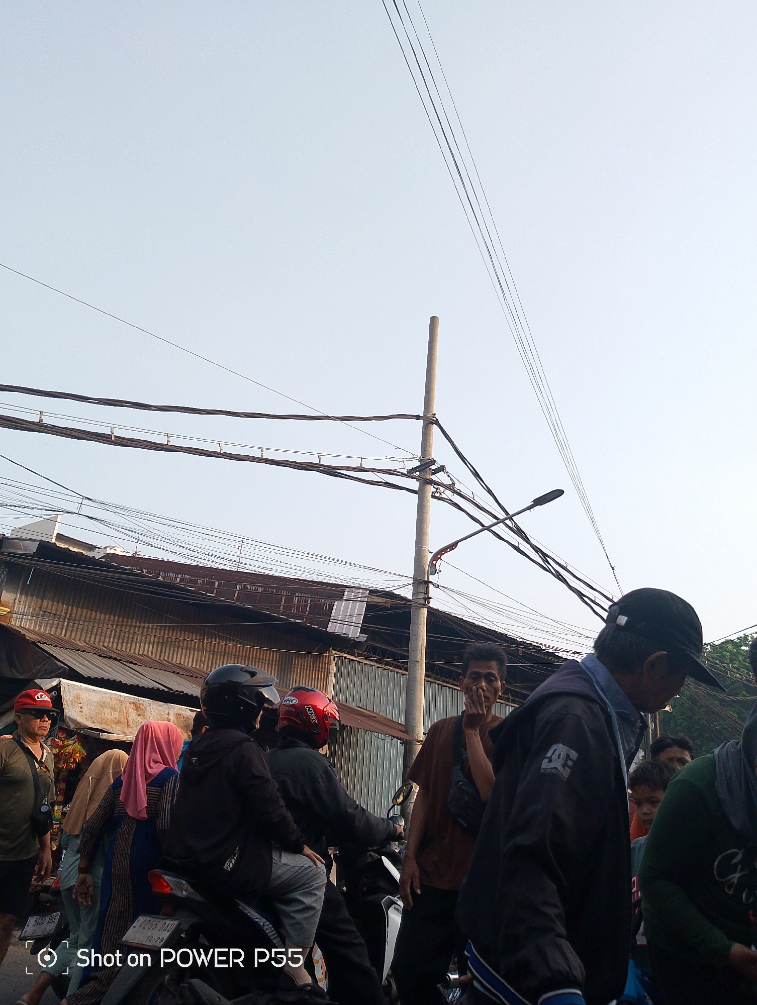 The image captures a street scene in Surabaya, Indonesia, on a clear day with a pale blue sky. A complex network of overhead electrical wires crisscrosses the frame, suspended from a tall, concrete utility pole. Below the wires, the corrugated metal roofs of buildings line the street, some showing signs of age.
In the foreground, a group of people are gathered. A man wearing a dark baseball cap and a black jacket with a prominent "DC Shoes" logo walks towards the right, his back mostly to the camera. To his left, another man with dark hair wears a brown t-shirt and has his hand near his face, looking towards the left.
Further to the left, a motorbike is partially visible, with two individuals on it. The person in the back wears a black helmet, and the person in front wears a red helmet. Beside them, several people are walking, including a woman in a pink headscarf, another woman in a light brown headscarf, and a man wearing a red cap and sunglasses. The overall atmosphere appears to be that of a busy urban environment.