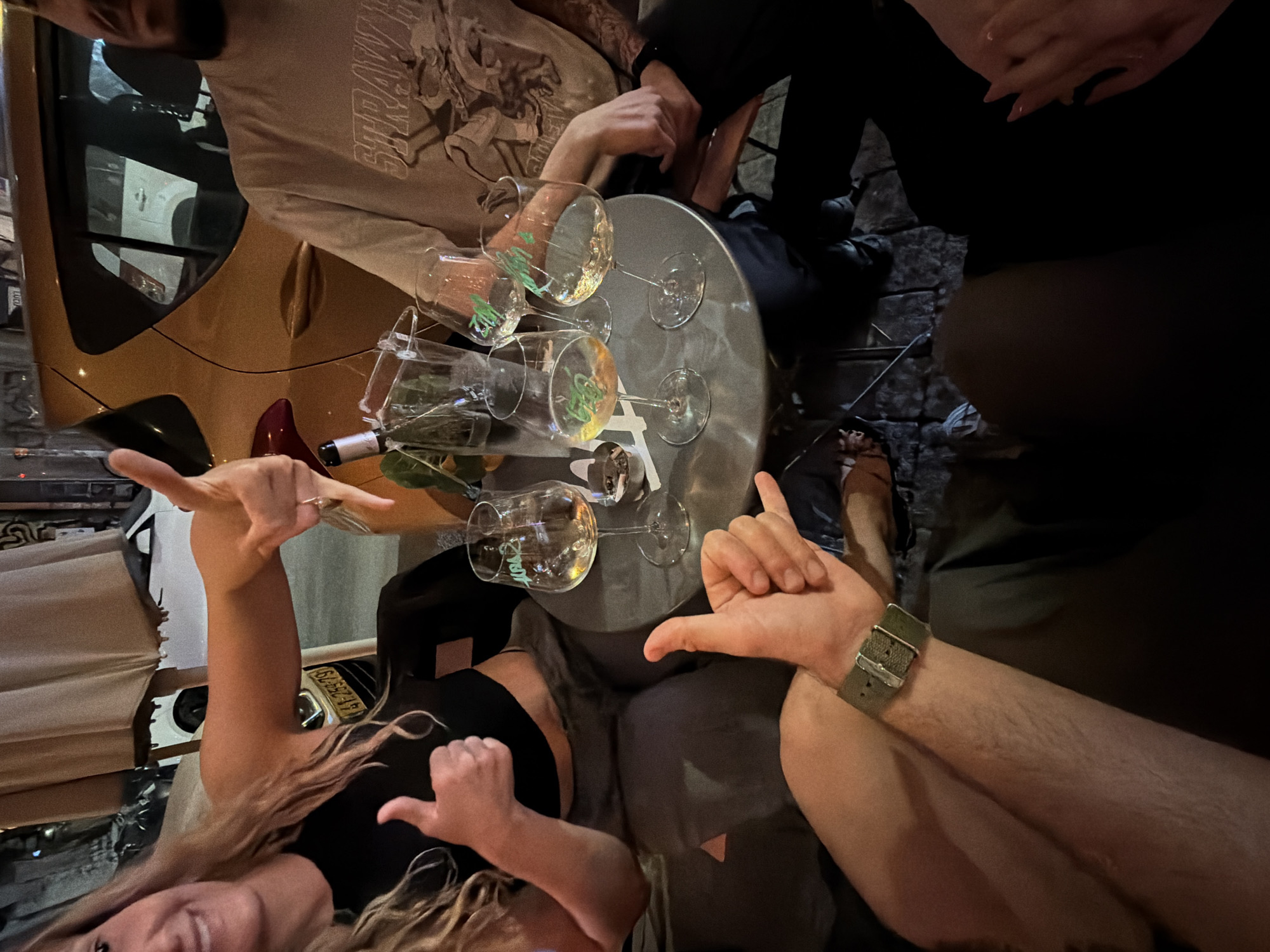 The image shows a group of people gathered around a small, round metal table outdoors in Tel-Aviv, Israel. Several wine glasses are on the table, some with liquid in them. A wine bottle also sits on the table. People are making the 'hang loose' hand gesture. A gold-colored car is visible in the background. The environment suggests a casual, social setting, possibly at a bar or restaurant. The setting suggests the scene occurred in the evening or night. The license plate is readable.