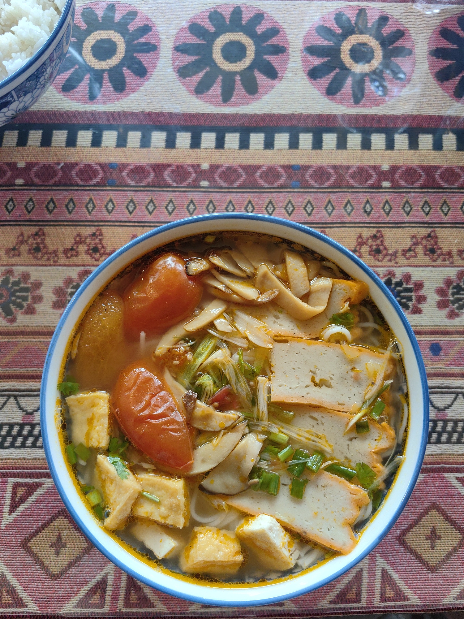The image shows a bowl of Vietnamese noodle soup on a patterned tablecloth. The soup contains tofu, tomatoes, mushrooms, and what appear to be fish cakes or tofu skins. Chopped green onions garnish the soup. In the upper left corner of the image, there is a bowl of white rice. The photo was taken in Thành phố Hà Giang, Vietnam.