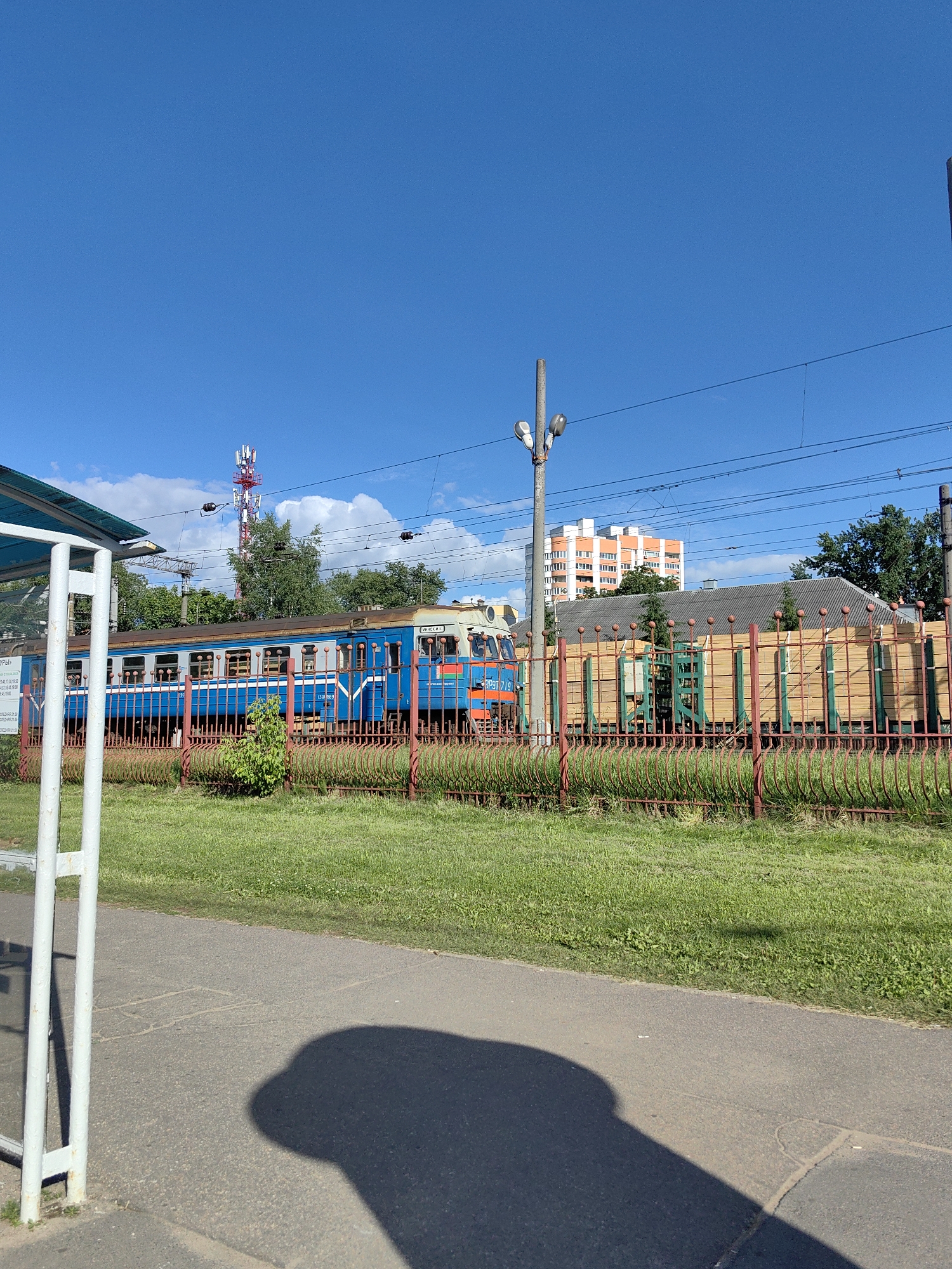 The image captures a sunny day scene at a train station or railway embankment in Minsk, Belarus. A blue and white train, likely a passenger train, is visible moving along the tracks, partially obscured by a red and green fence. Behind the fence, a grassy area borders the railway. In the background, there are trees, a modern orange-and-white apartment building, and a red-and-white telecommunications tower, indicating an urban or suburban setting.
The sky is a vibrant blue with a few scattered white clouds, suggesting pleasant weather and likely daytime. The lighting indicates it's mid-day or afternoon, with strong shadows cast on the asphalt path in the foreground. The shadow in the foreground appears to be of a person, partially obscuring a section of the path.
There is some visible text on a sign behind a white support pole in the lower left of the image, but it is too blurry to read. Another piece of text, "139 719", is visible on the side of the train. The overall impression is one of an ordinary day at a functional transportation hub, with the bright sunlight adding a cheerful tone to the scene.