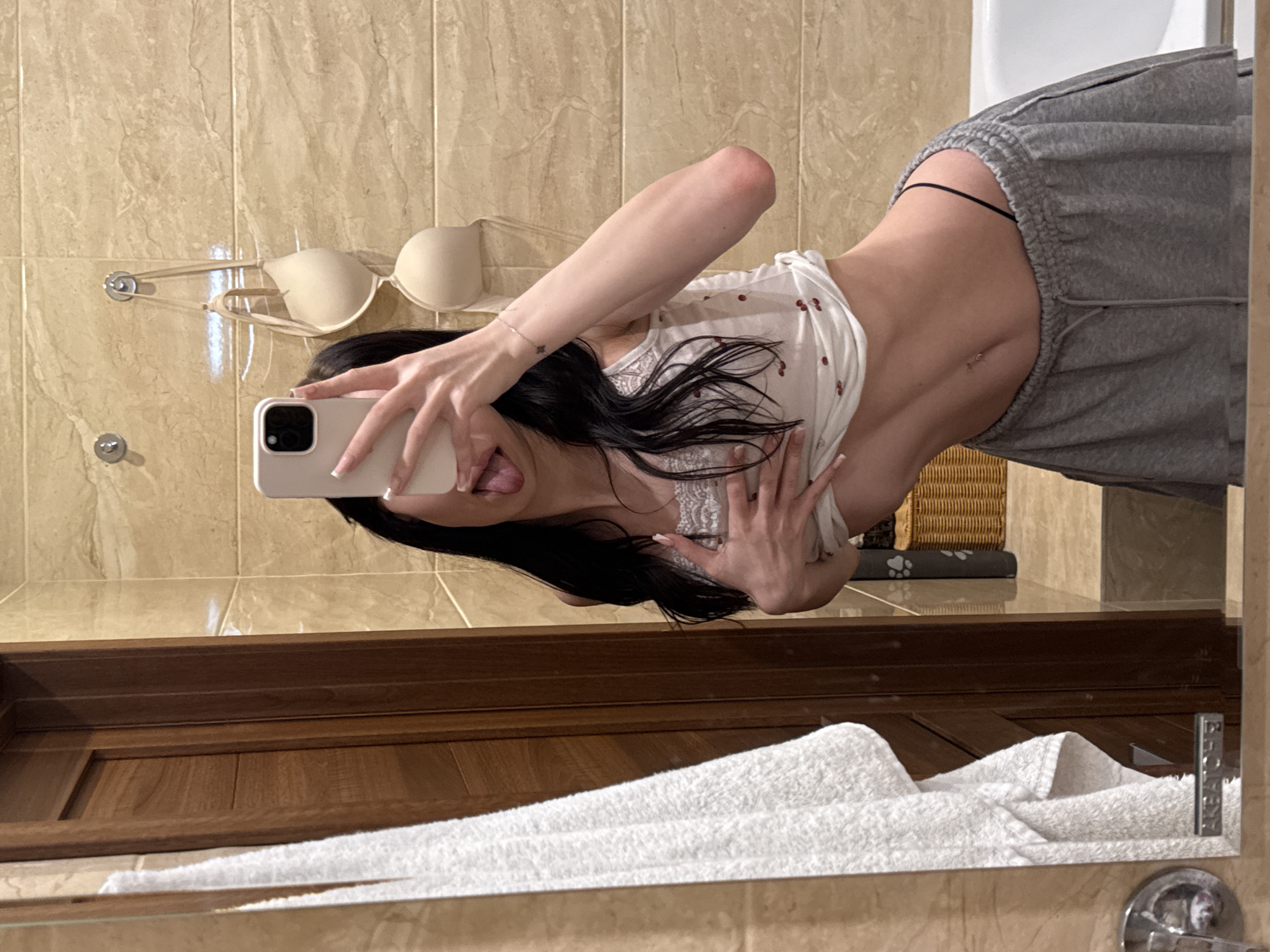 In a bathroom with beige tiled walls and a wooden door, a person is reflected in a mirror. The person has long black hair and is wearing a white tank top with a cherry pattern and grey sweatpants. They are holding their phone to take a selfie, with their tongue sticking out. Two beige bras are hanging on hooks on the wall behind them. A white towel is draped over a rack on the left side of the mirror. The scene appears to be in a private residence.