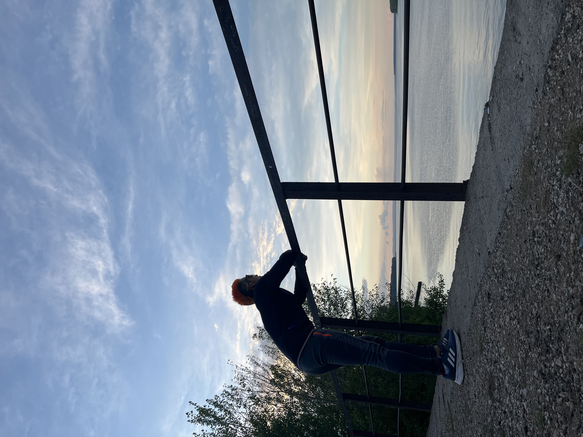 A man with orange hair performs a pull-up on a black metal bar overlooking a body of water. The sky is visible with light clouds. The man is wearing a black long-sleeved shirt, gray pants, and blue sneakers with white stripes. The location appears to be outdoors, possibly a park or scenic overlook. The text indicates the city is Chaikovsky, Russia. The ground is covered in gravel. Trees and other vegetation are visible below the bar.