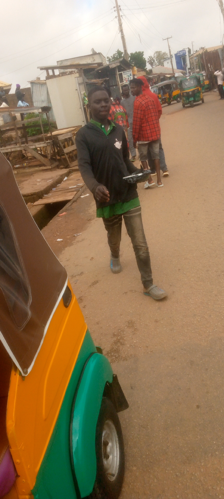 A young man walks down a dirt road, holding a black rectangular object in his right hand. He is wearing a black long-sleeved shirt with a green collar and a graphic on the left chest. His pants are dark and appear to be ripped or distressed. He is wearing grey clogs on his feet. In the foreground to the left, a portion of a yellow and green auto-rickshaw is visible. Behind the man, other individuals are present, along with market stalls and buildings. Several other auto-rickshaws are parked or driving in the background. The sky is overcast.